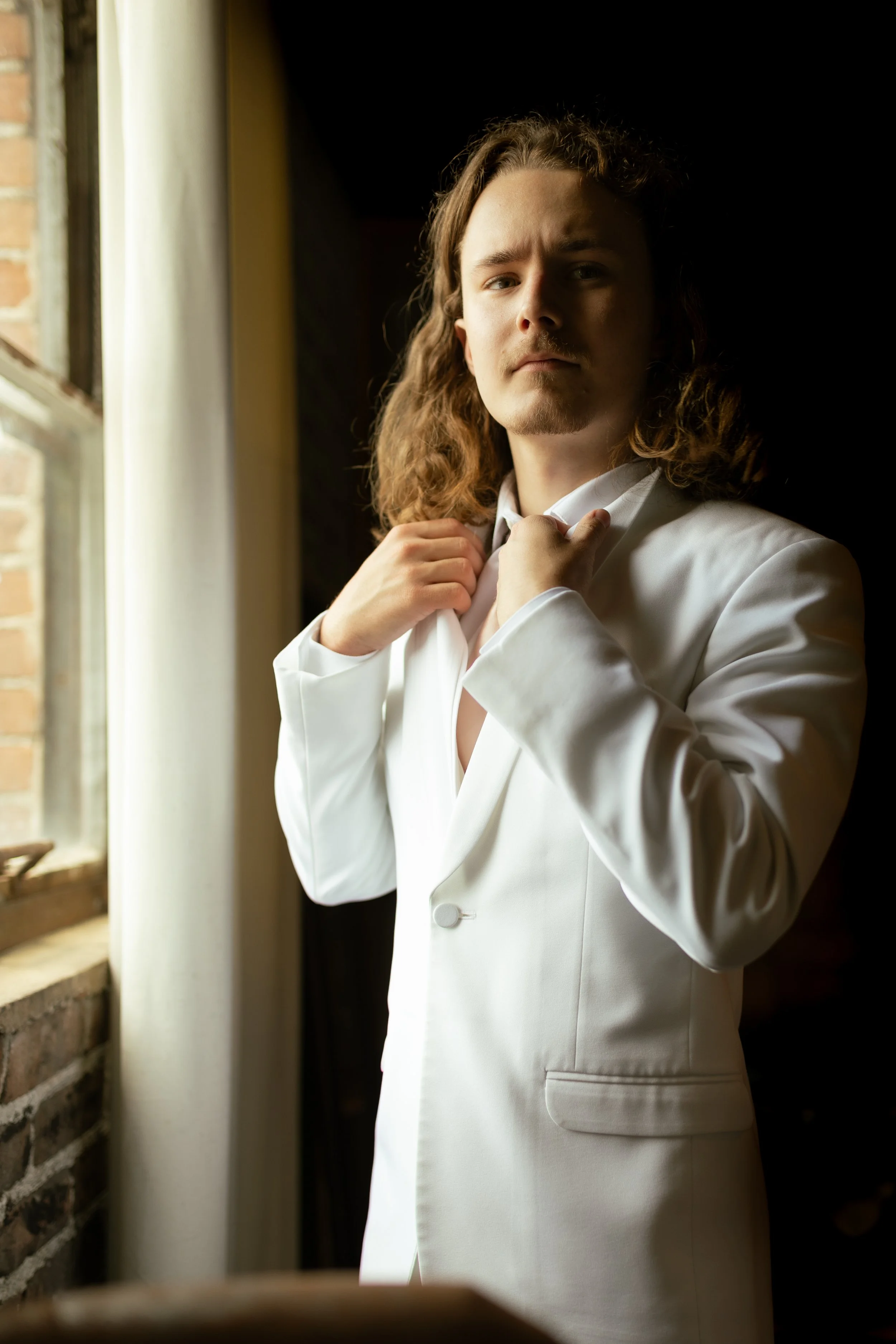 A man with long, curly hair standing by a window, adjusting his white suit jacket, with indoor lighting and brick wall visible. Wedding Photography at The Venue in Hattiesburg, MS.
