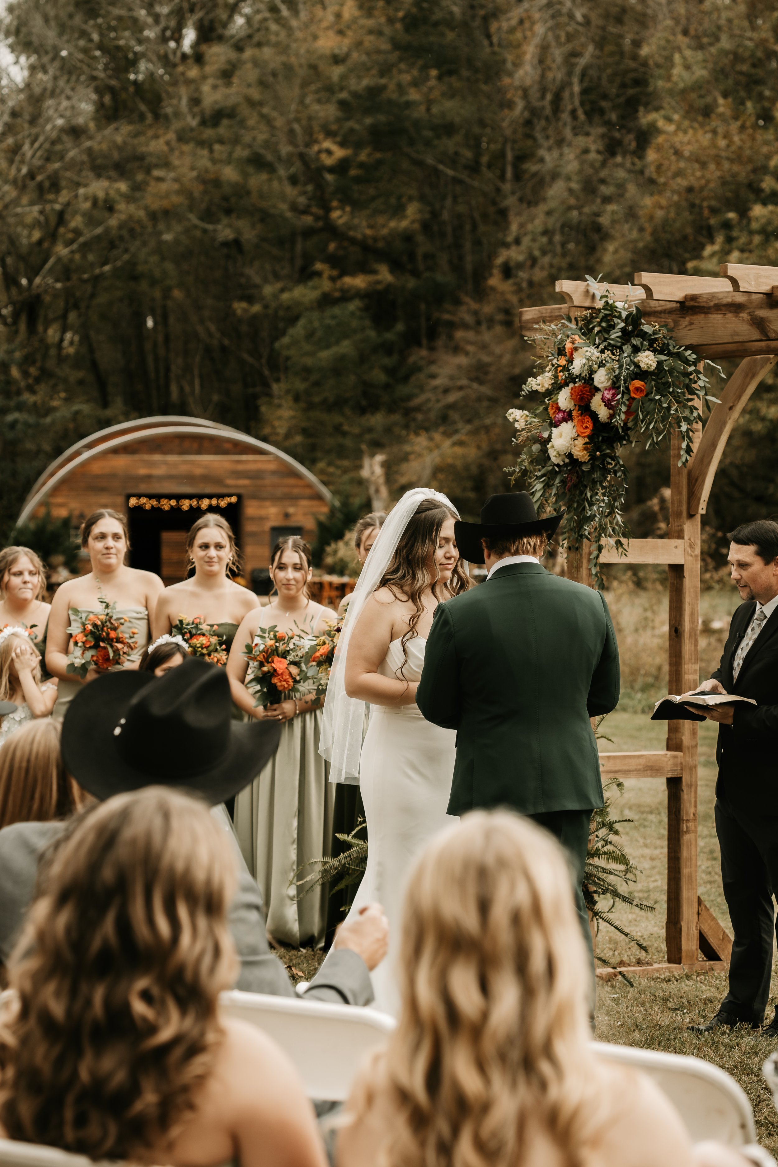 A wedding ceremony outdoors with the bride and groom exchanging vows, surrounded by bridesmaids, guests, and a decorated wooden arch, in a natural setting with trees and a wooden structure in the background.