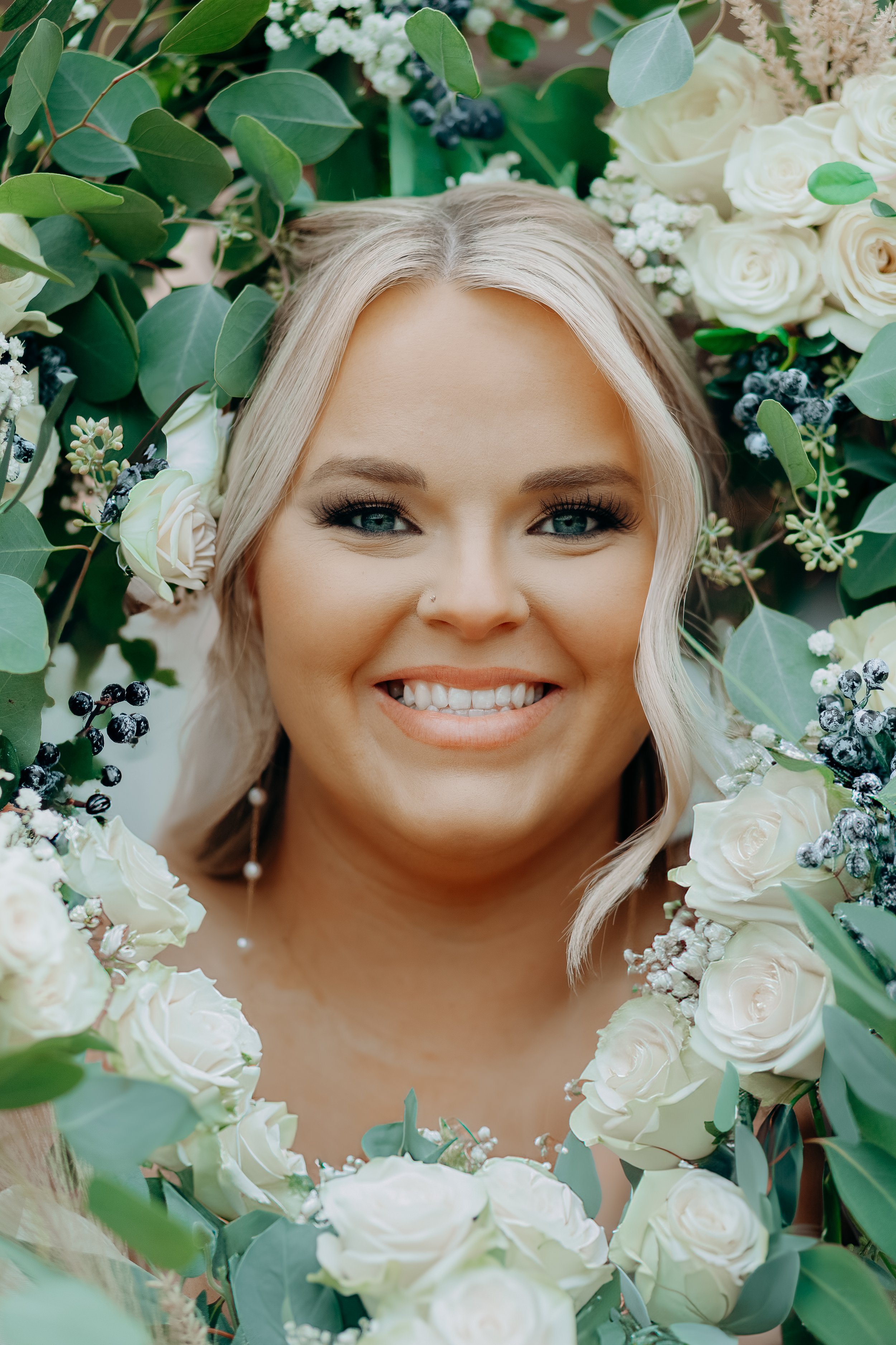 A woman with blonde hair and blue eyes smiling among white roses, greenery, and berries.