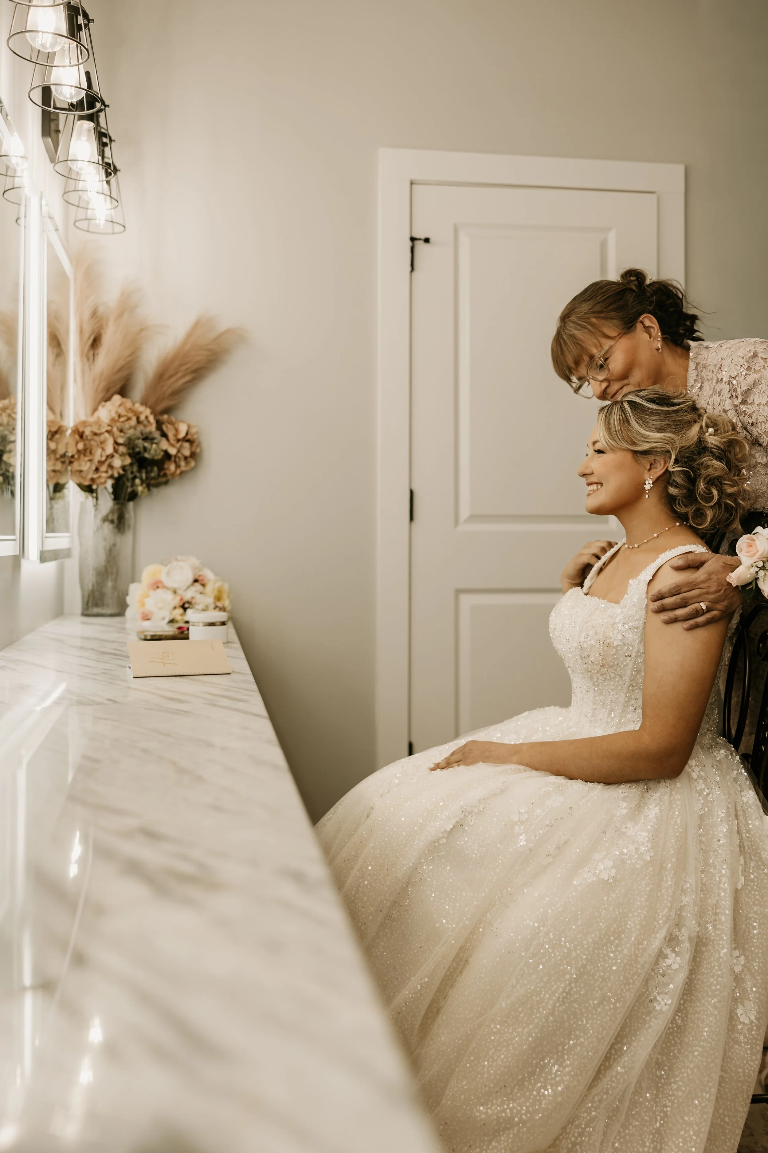 Bride and Mother sitting at vanity as mother of the bride looks and admires her daughter. The Bride gazes at herself in the mirror.