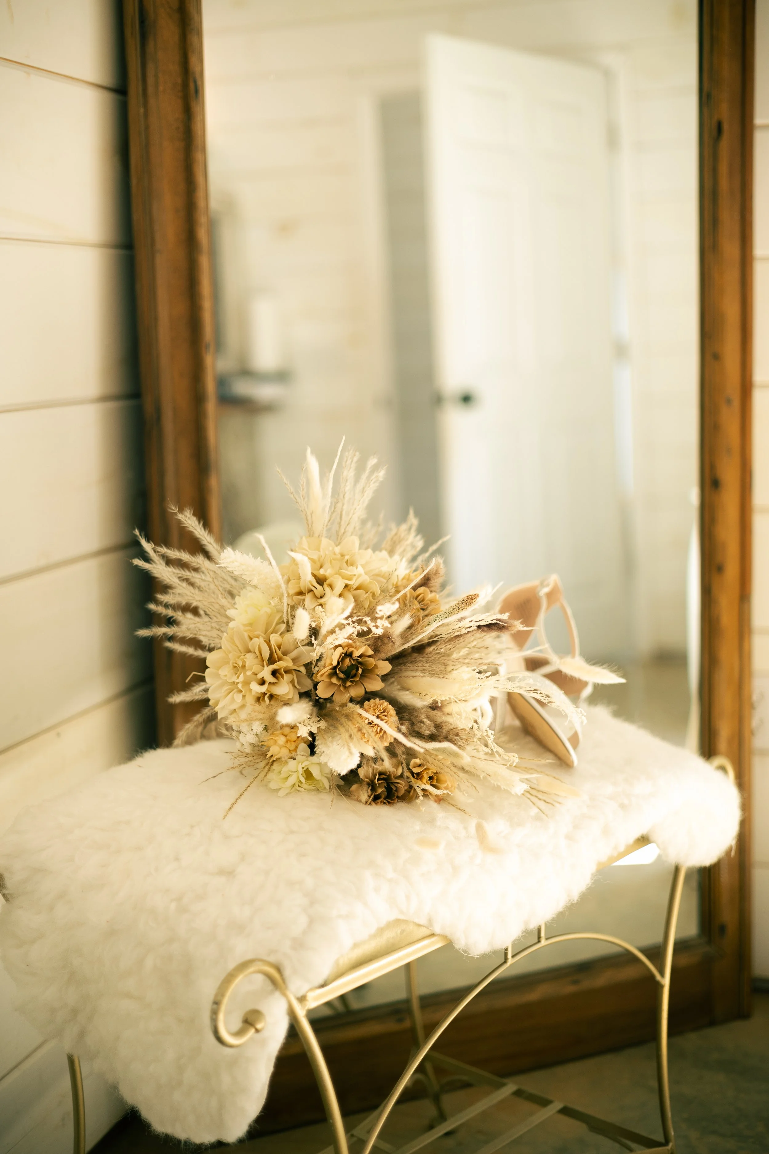 A bouquet of dried flowers and pampas grass resting on a white sheepskin-covered bench with a full-length mirror and a door in the background. Wedding photographer in Mississippi.