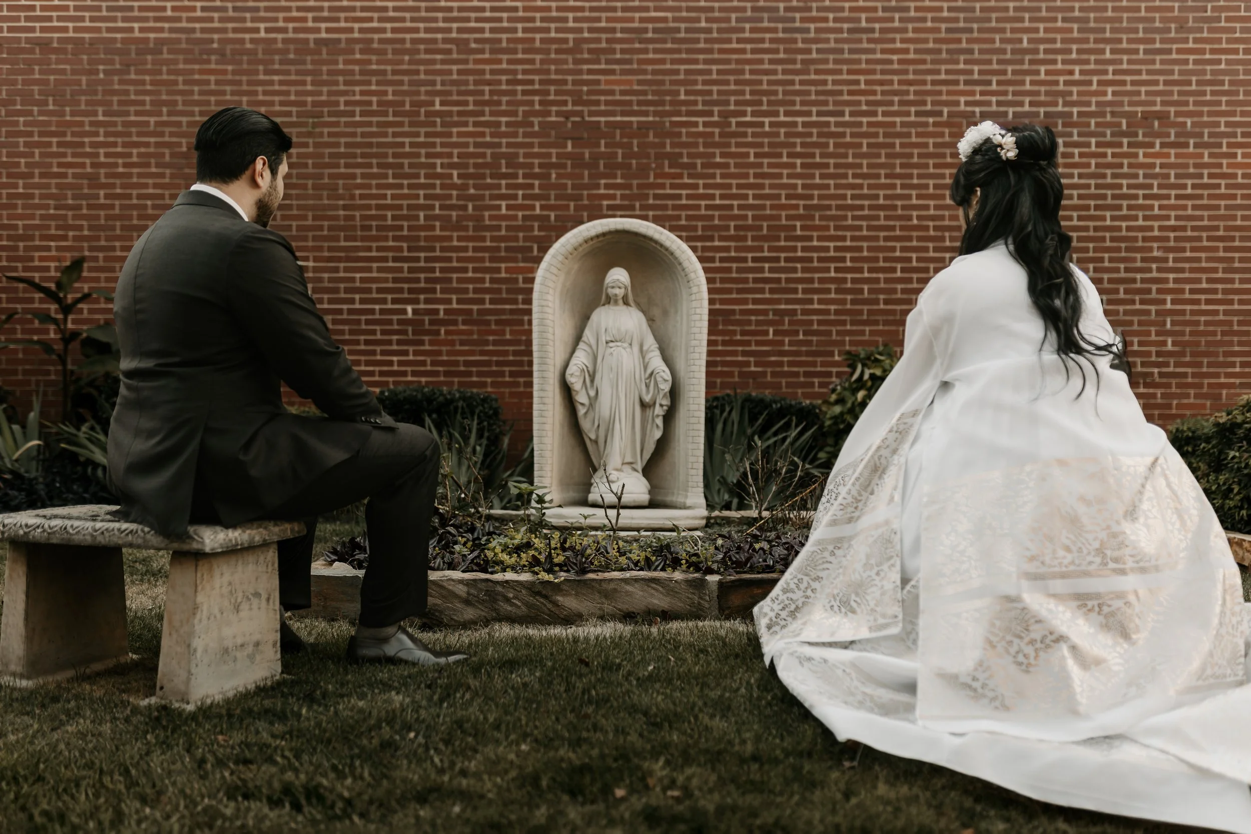 A man and a woman in formal attire kneel before a religious statue of the Virgin Mary in a garden with a brick wall background.