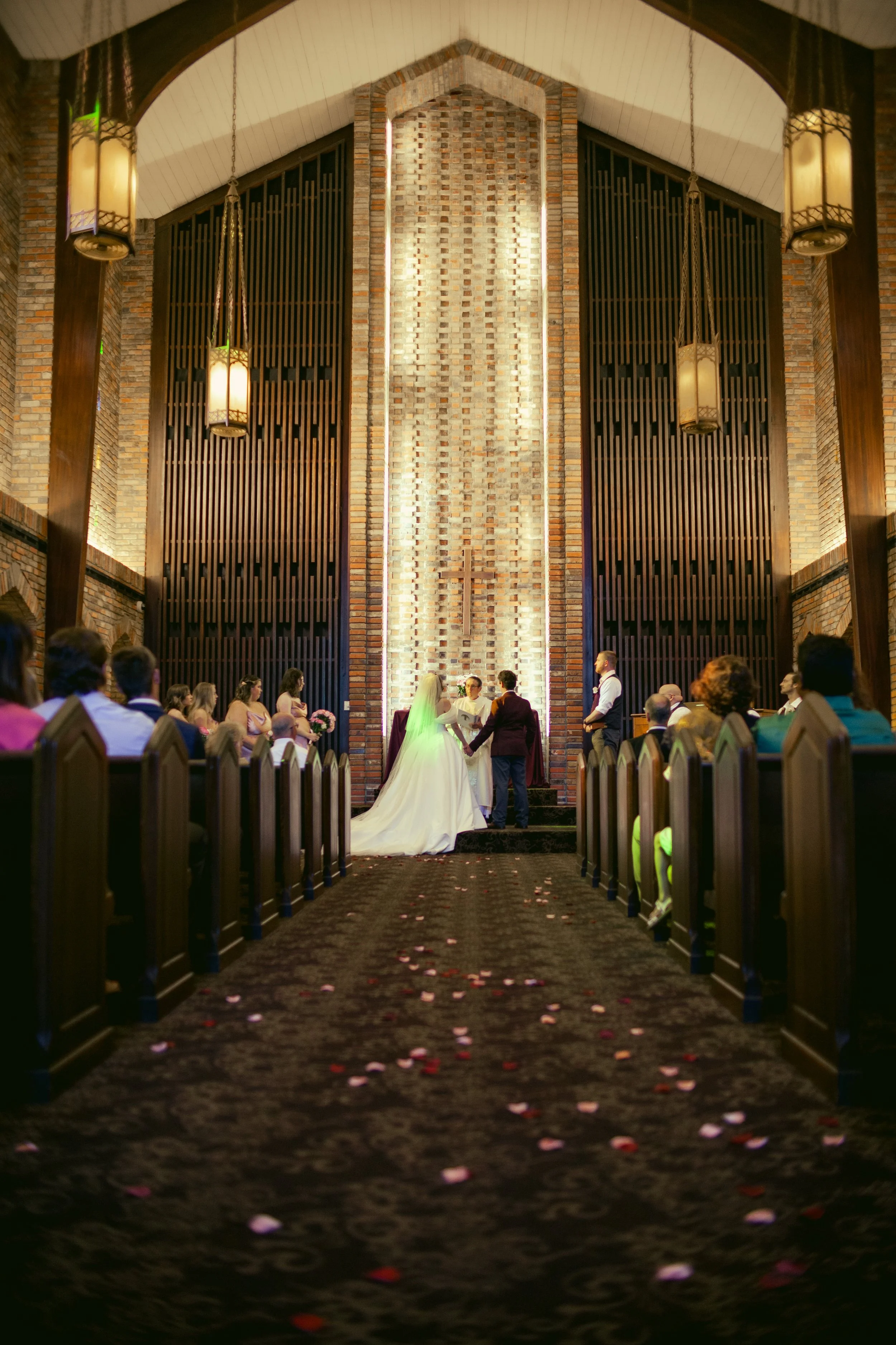 A wedding ceremony inside a church with a bride and groom standing at the altar, surrounded by guests seated in wooden pews, with rose petal confetti on the carpeted aisle.Chapel of Memories in Starkville, MS on a weding day.