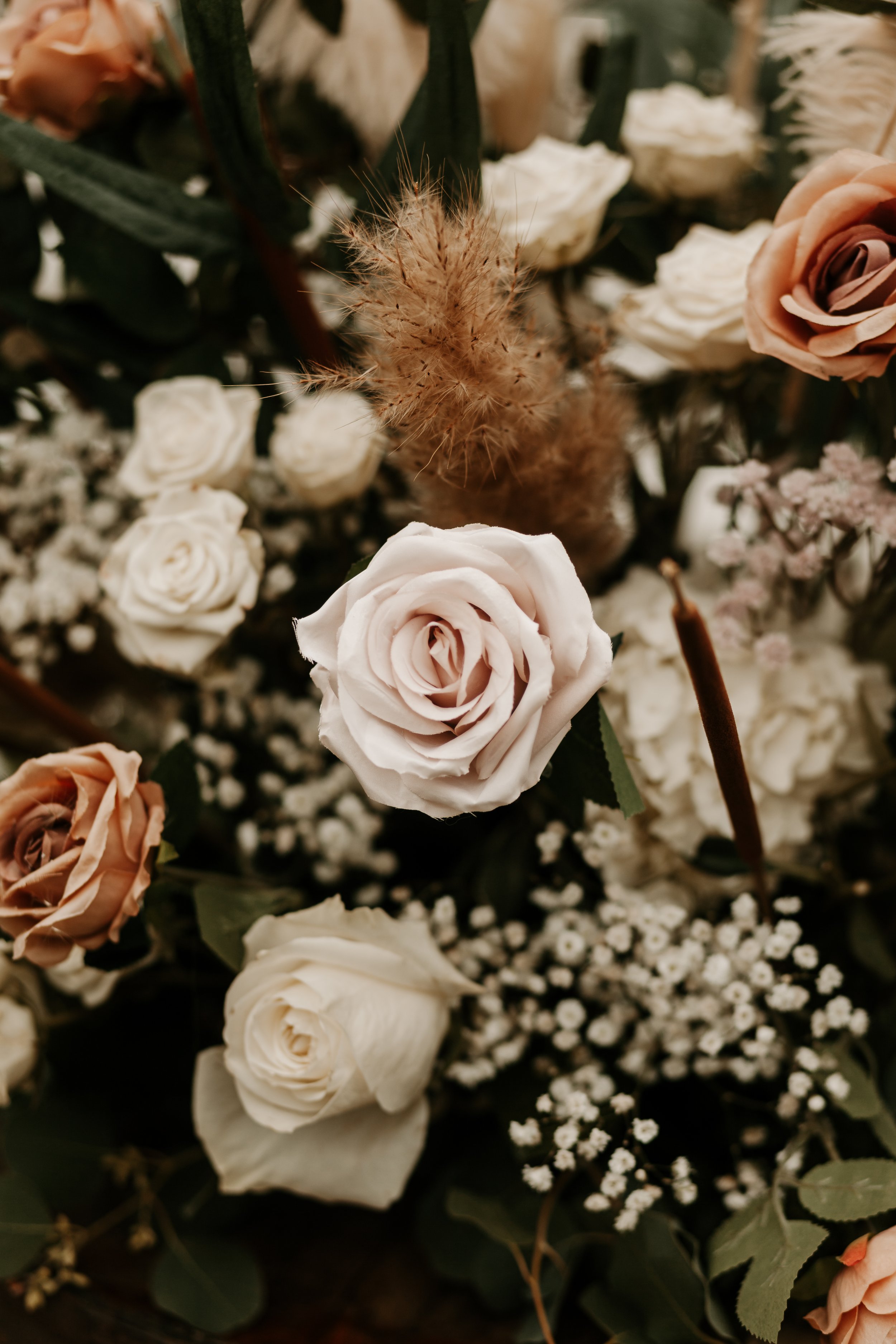 Close-up of a floral arrangement featuring white, cream, and blush roses, small white filler flowers, and dried brown flowers. Behind The Pines Wedding Venue. Wedding Photography in Mississippi.