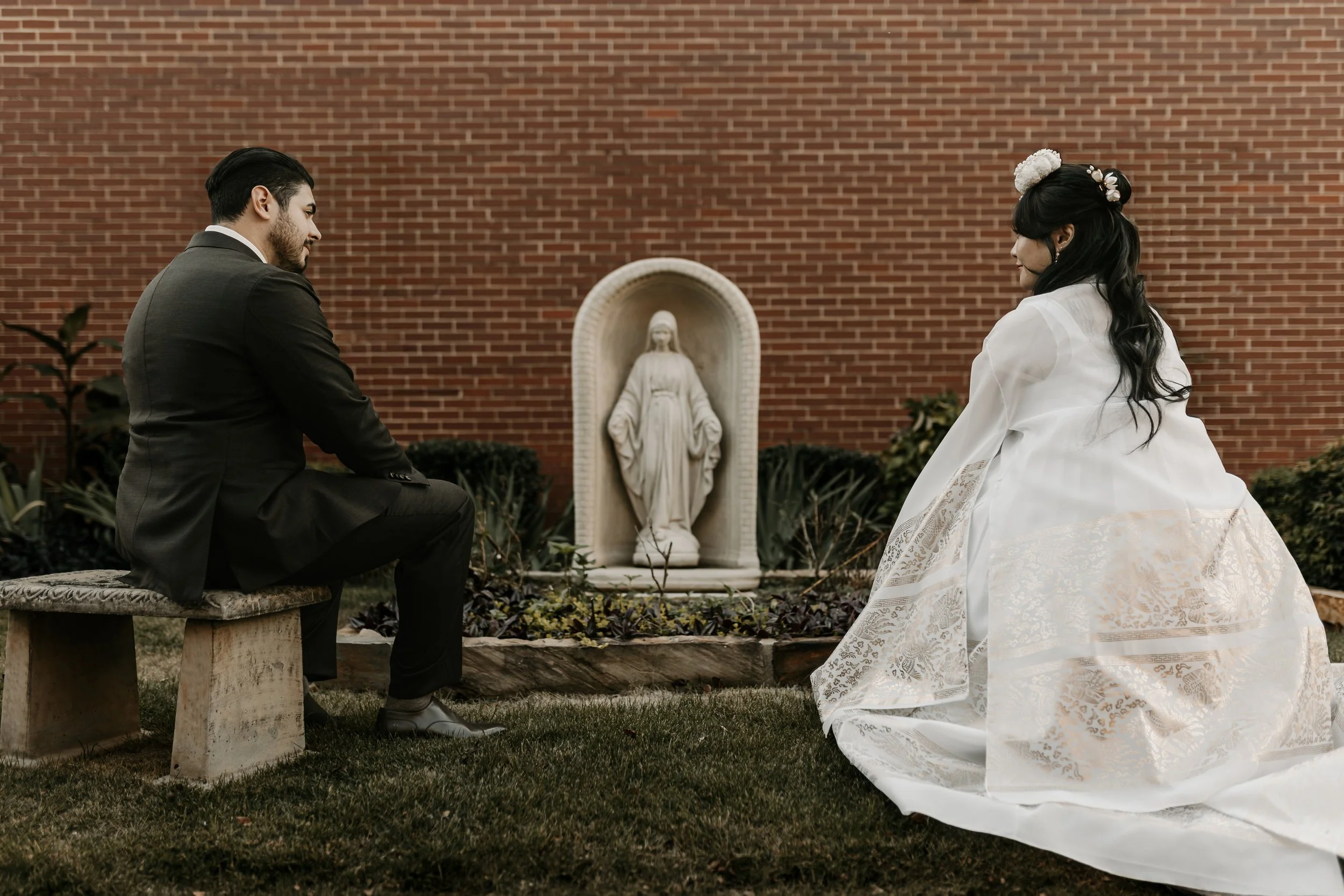 A man in a black suit and a woman in a white wedding dress kneeling outdoors near a religious statue of Jesus, with a brick wall in the background.