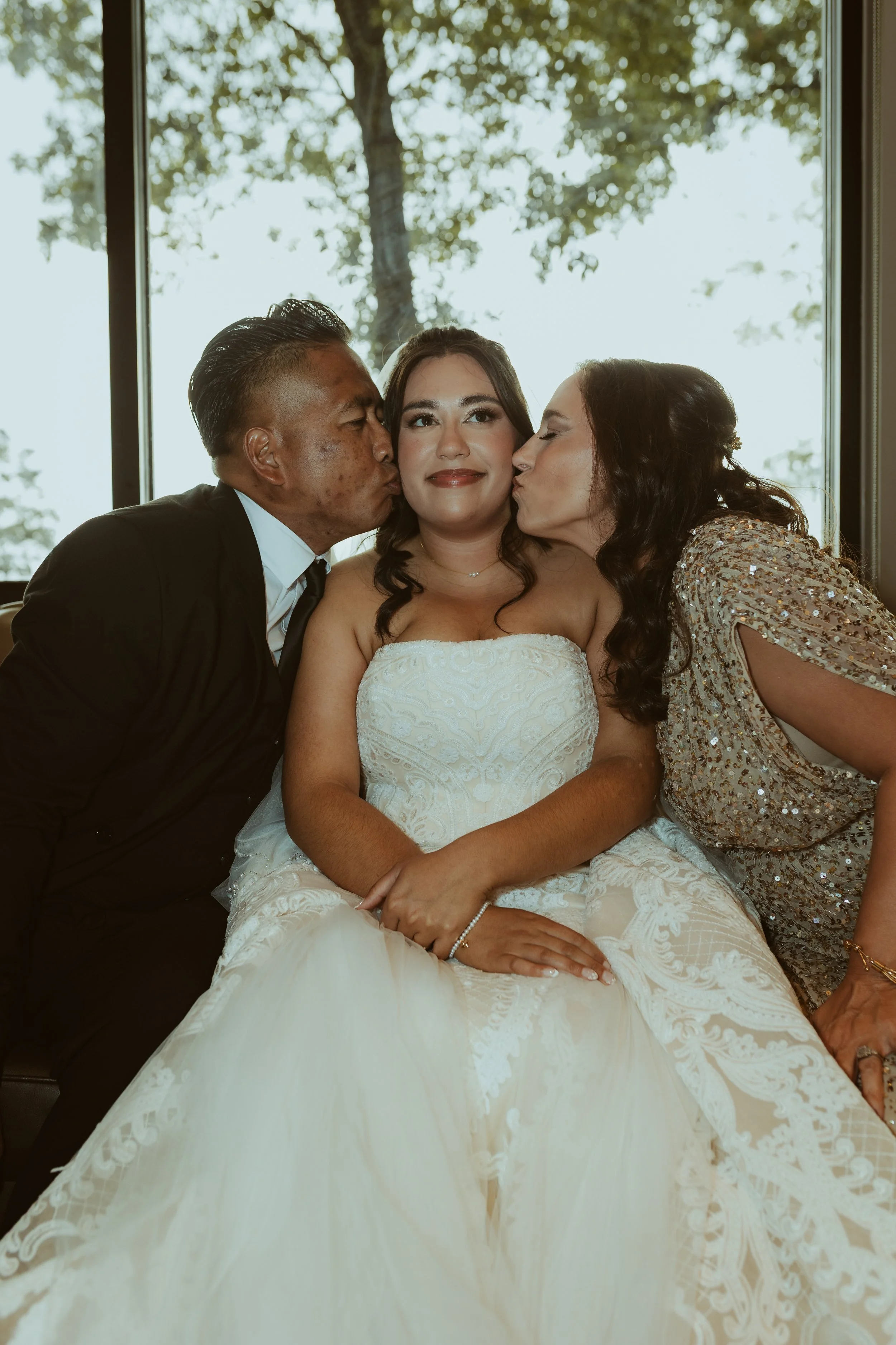 A woman in a white wedding dress sitting between a man in a black suit and a woman in a sparkly silver dress, with both giving her kisses on the cheeks. They are inside, with a large window revealing trees outside, and natural light illuminating thei
