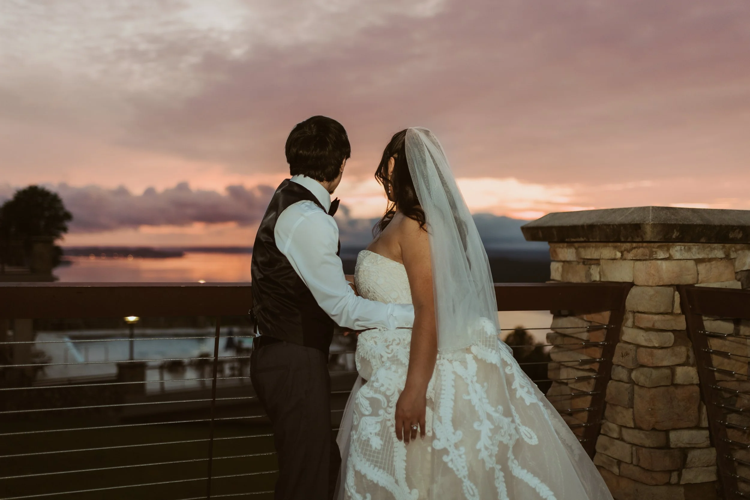 A couple dressed in wedding attire stands on a terrace at sunset, with a lake and cloudy sky in the background. Guntersville, AL Weddings. Wedding photographer in guntersville, al.