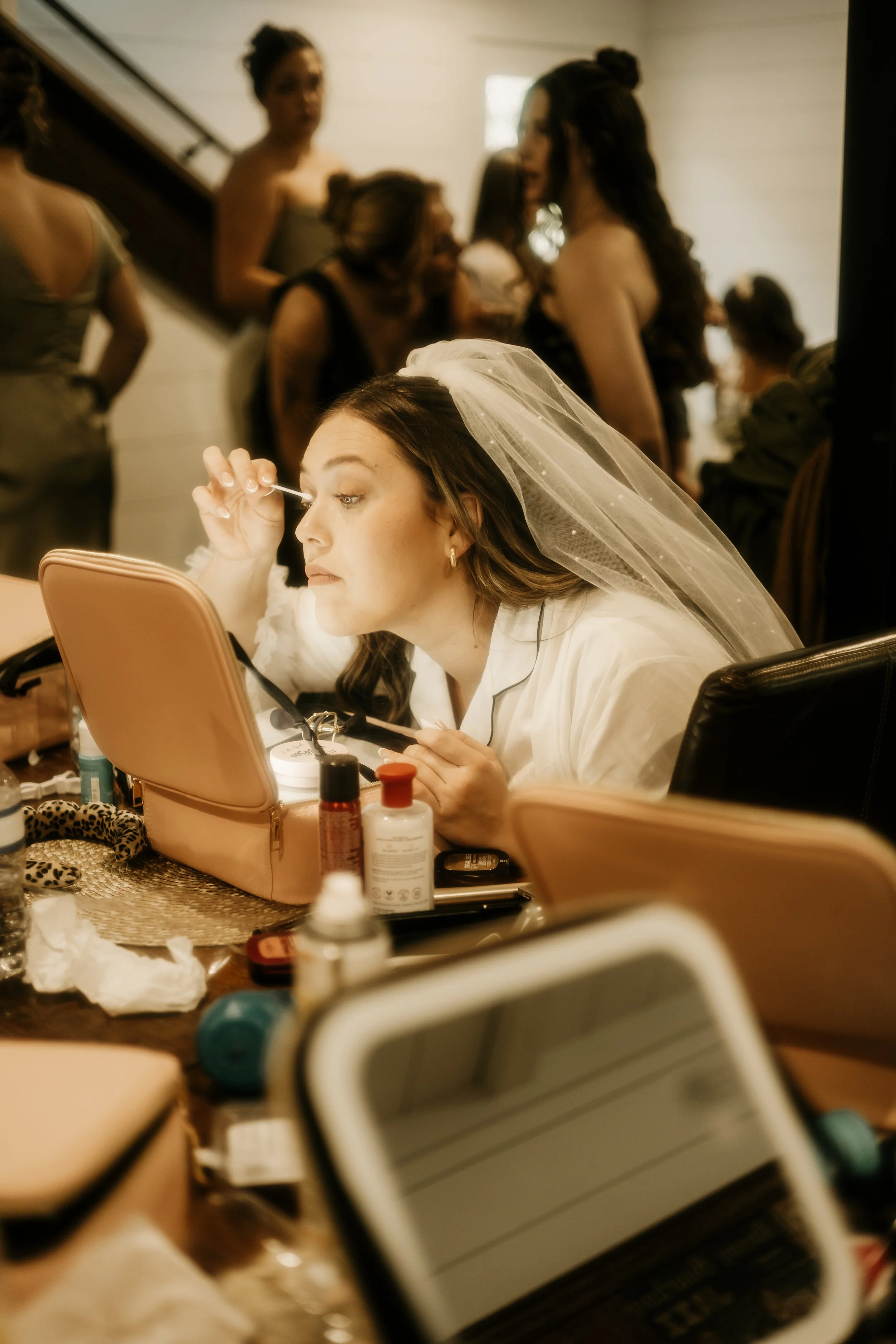 Bride in a white gown and veil applying makeup at a vanity mirror surrounded by makeup items, with bridesmaids in casual dresses in the background. Story telling wedding photography in Mississippi.