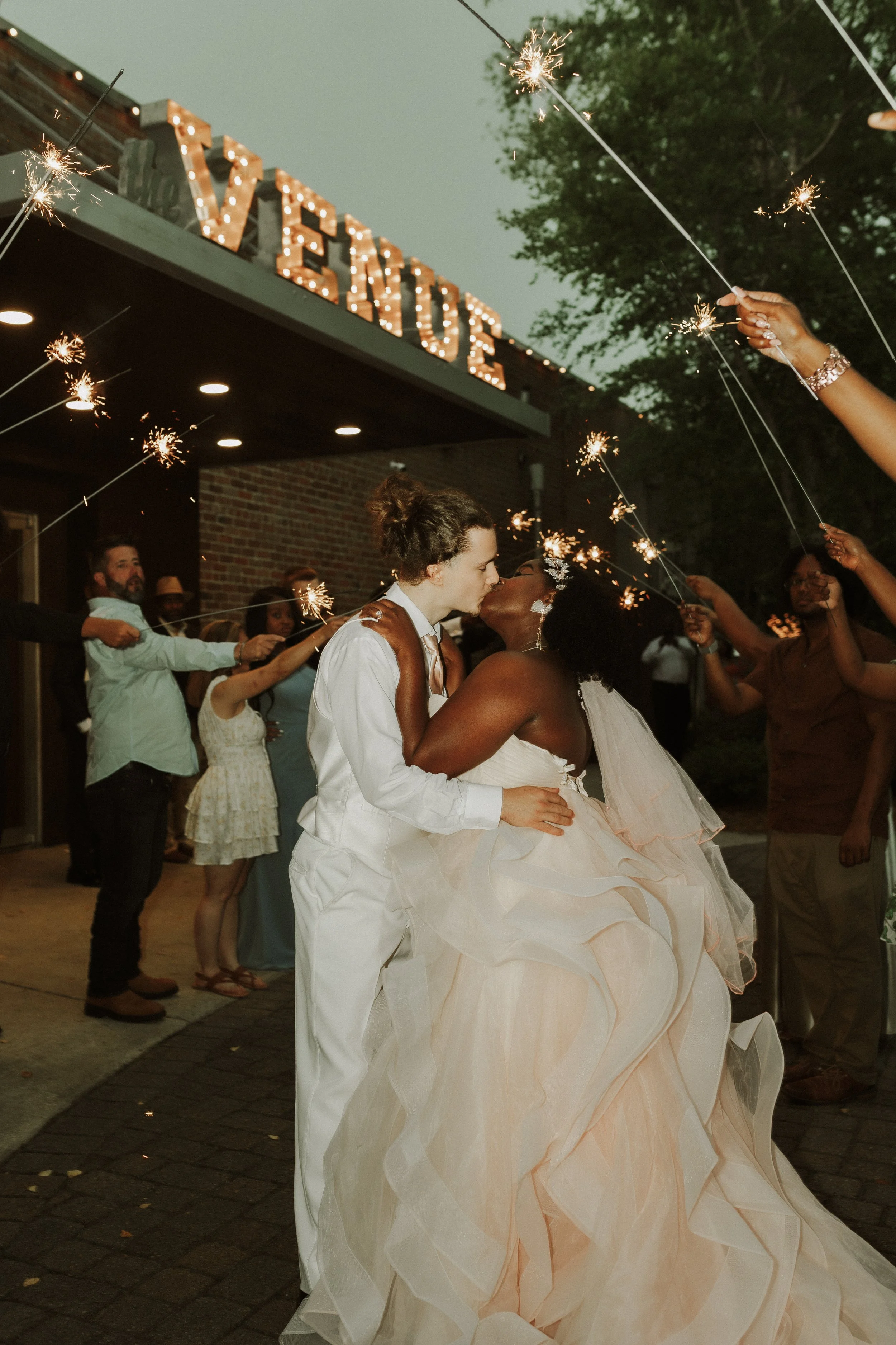 Two brides, one with curly black hair and the other with brown hair, sharing a kiss at their wedding celebration. They are surrounded by guests holding sparklers, with a lit sign that reads 'LOVE' above them.