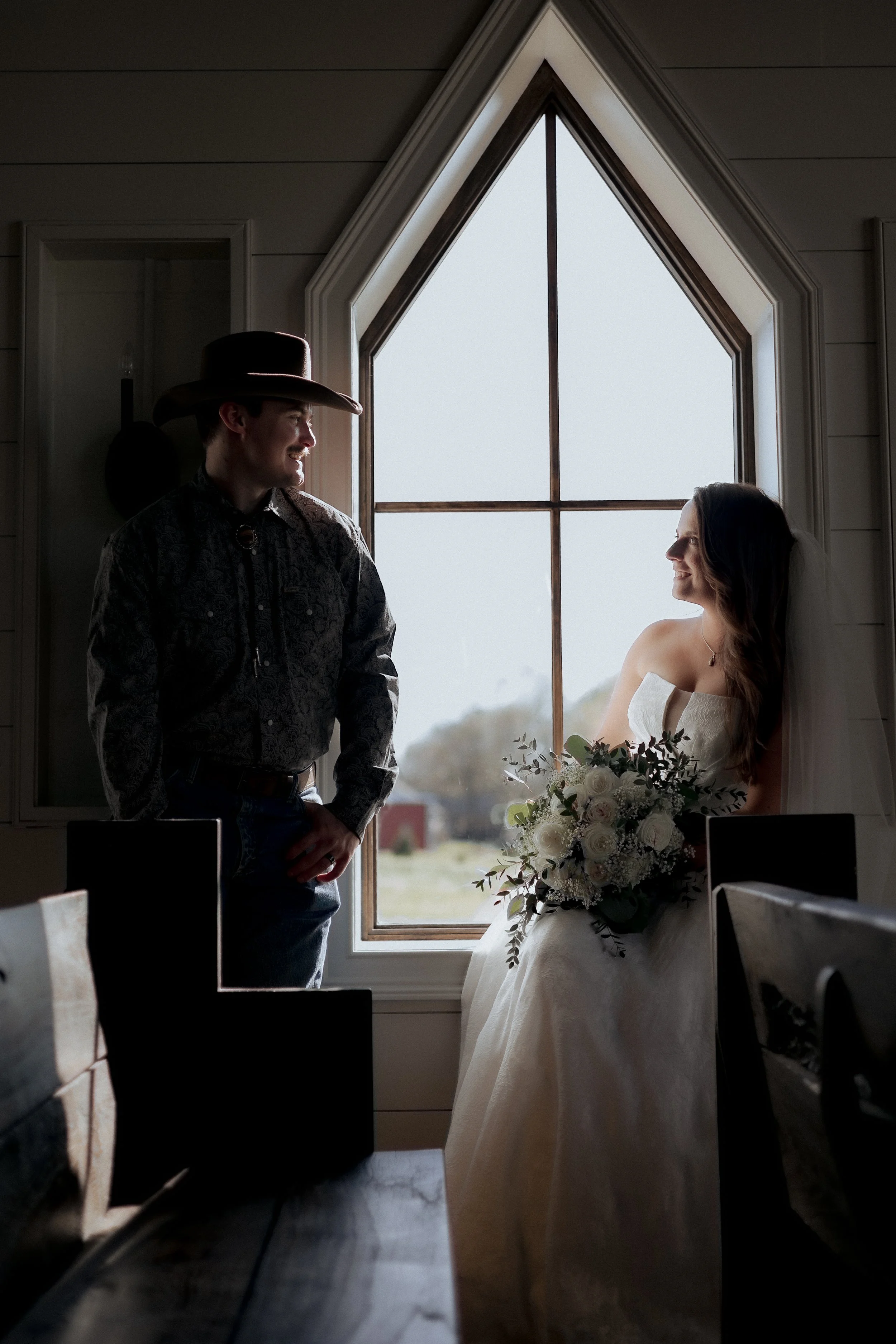 A bride and groom are standing inside a room with a large window. The groom is wearing a cowboy hat and dark shirt, while the bride is holding a large bouquet of white flowers and looking at him, both smiling. Taken at Bainfield Farm Venue.
