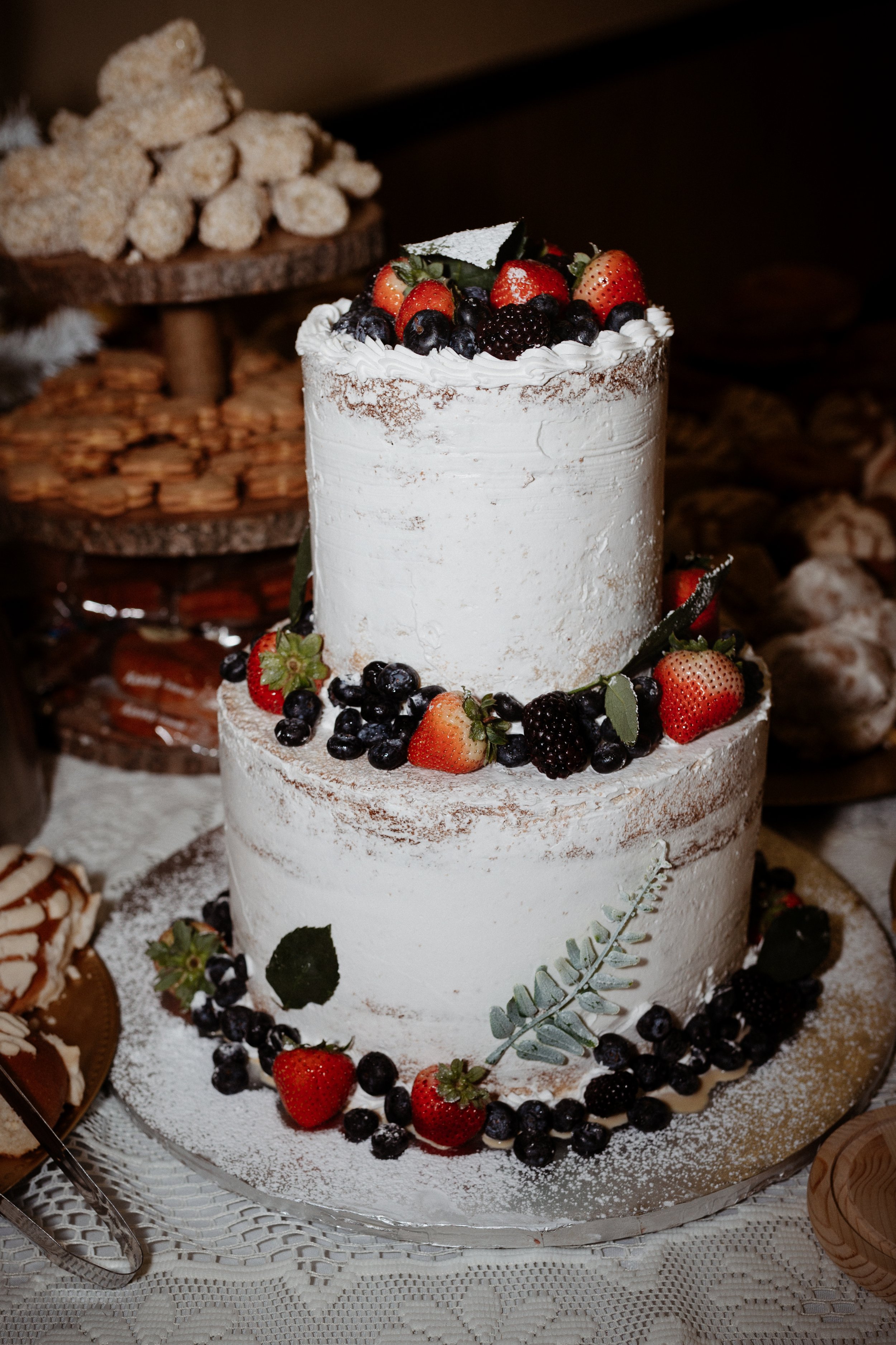 Two-tiered white frosted cake decorated with fresh strawberries, blueberries, blackberries, and green leaves, with powdered sugar on the base and top. The cake is on a glass plate surrounded by additional berries and leaves.