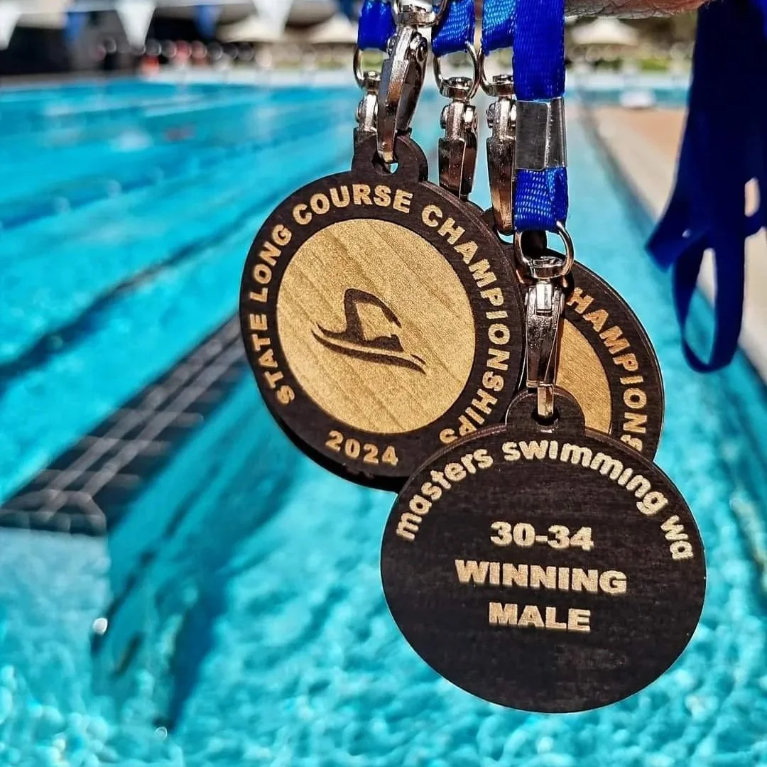 Swimmer medals hanging on a blue ribbon with a pool in the background, celebrating success at the 2024 State Long Course Championship for 30-34 age male winners.