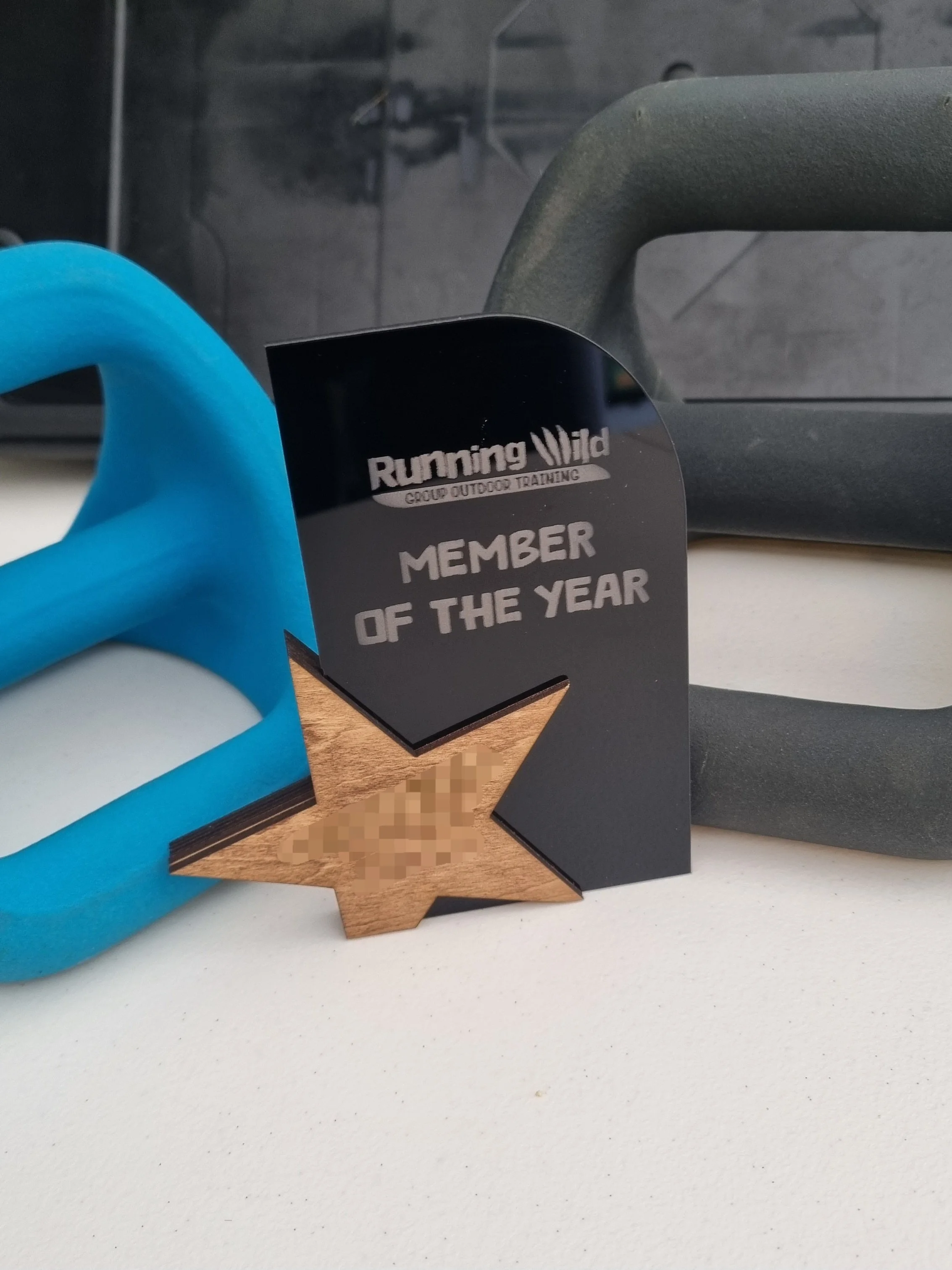 Award badge reading 'Member of the Year' from Running Wild Group Outdoor Training, with a wooden star, on a white table with gym equipment in the background.