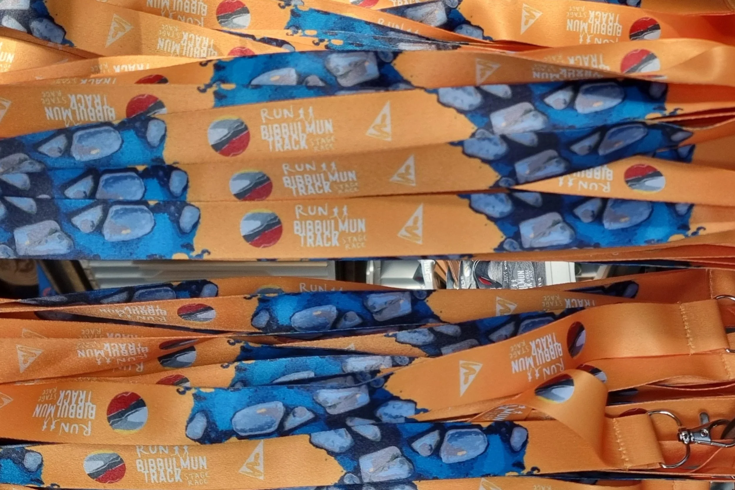 Multiple orange lanyards with blue and gray rocks pattern and text "Run Bibbulmun Track Stage Race" displayed, arranged in rows.