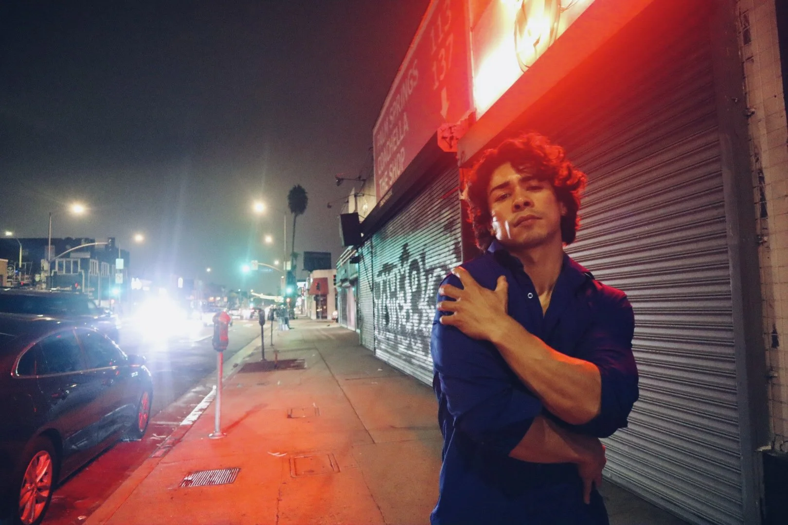 A young man with curly hair wearing a dark shirt stands on a city sidewalk at night, arms crossed over his chest, with a closed storefront and streetlights in the background.