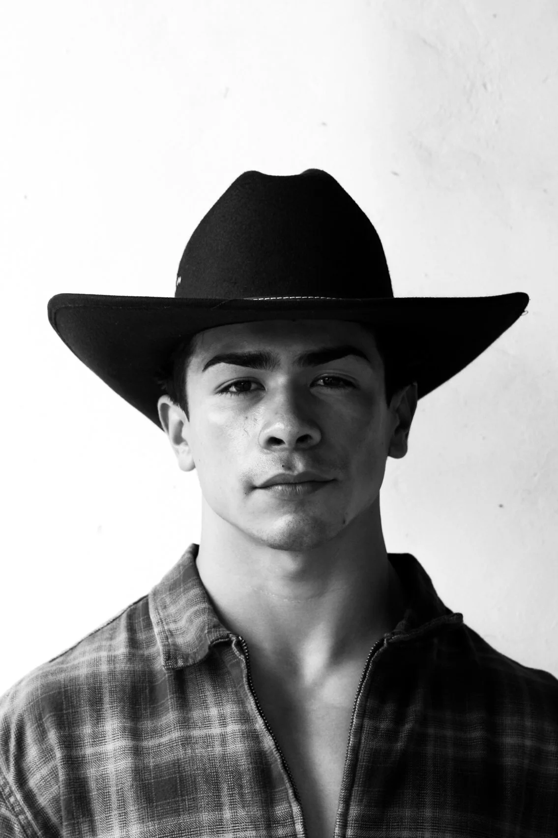 Black and white portrait of a young man wearing a wide-brimmed cowboy hat and a checkered shirt, looking directly at the camera with a slight smile, against a plain background.
