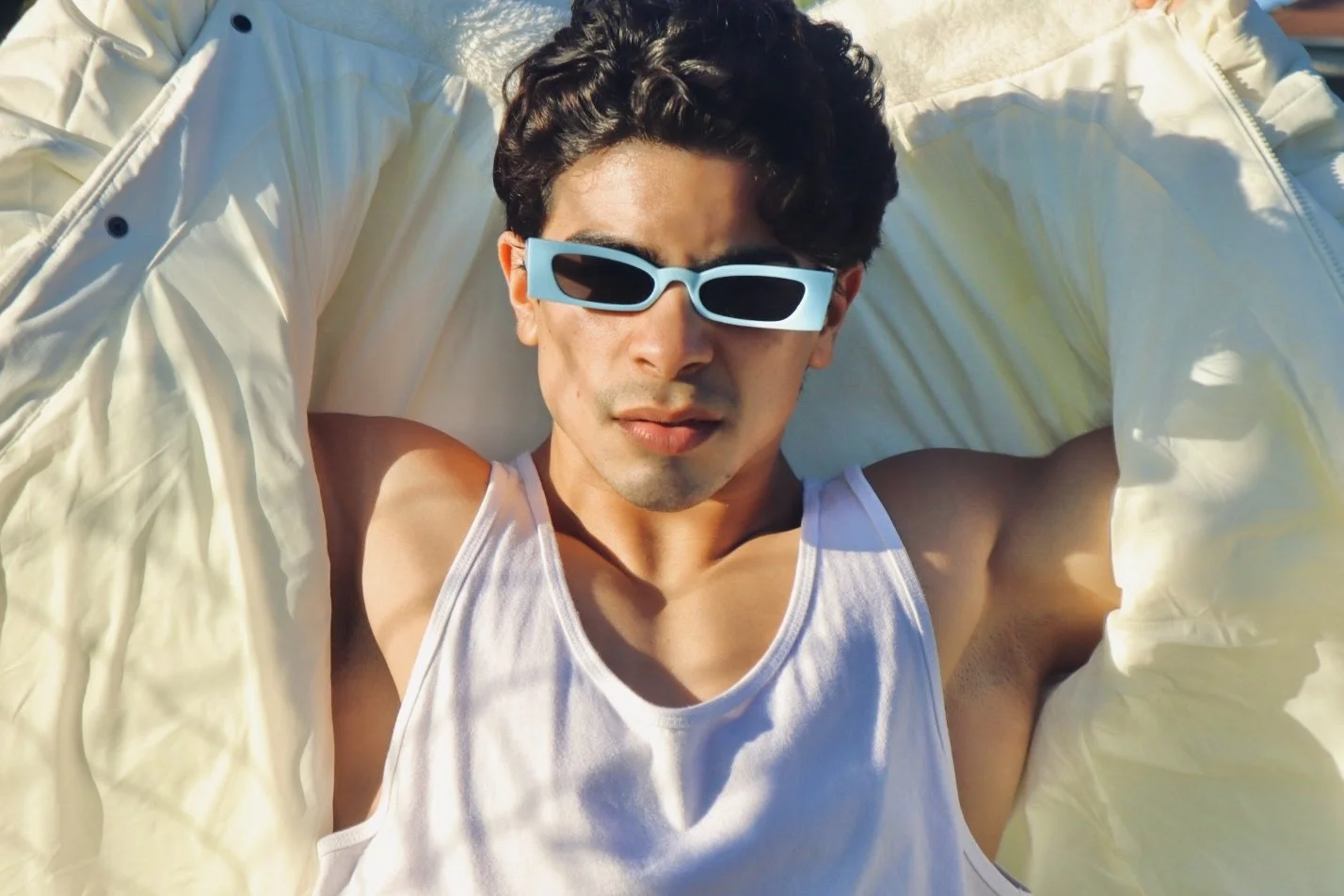Young man lying on a white pillow outdoors, wearing a white tank top and sunglasses, relaxed and enjoying sunlight.