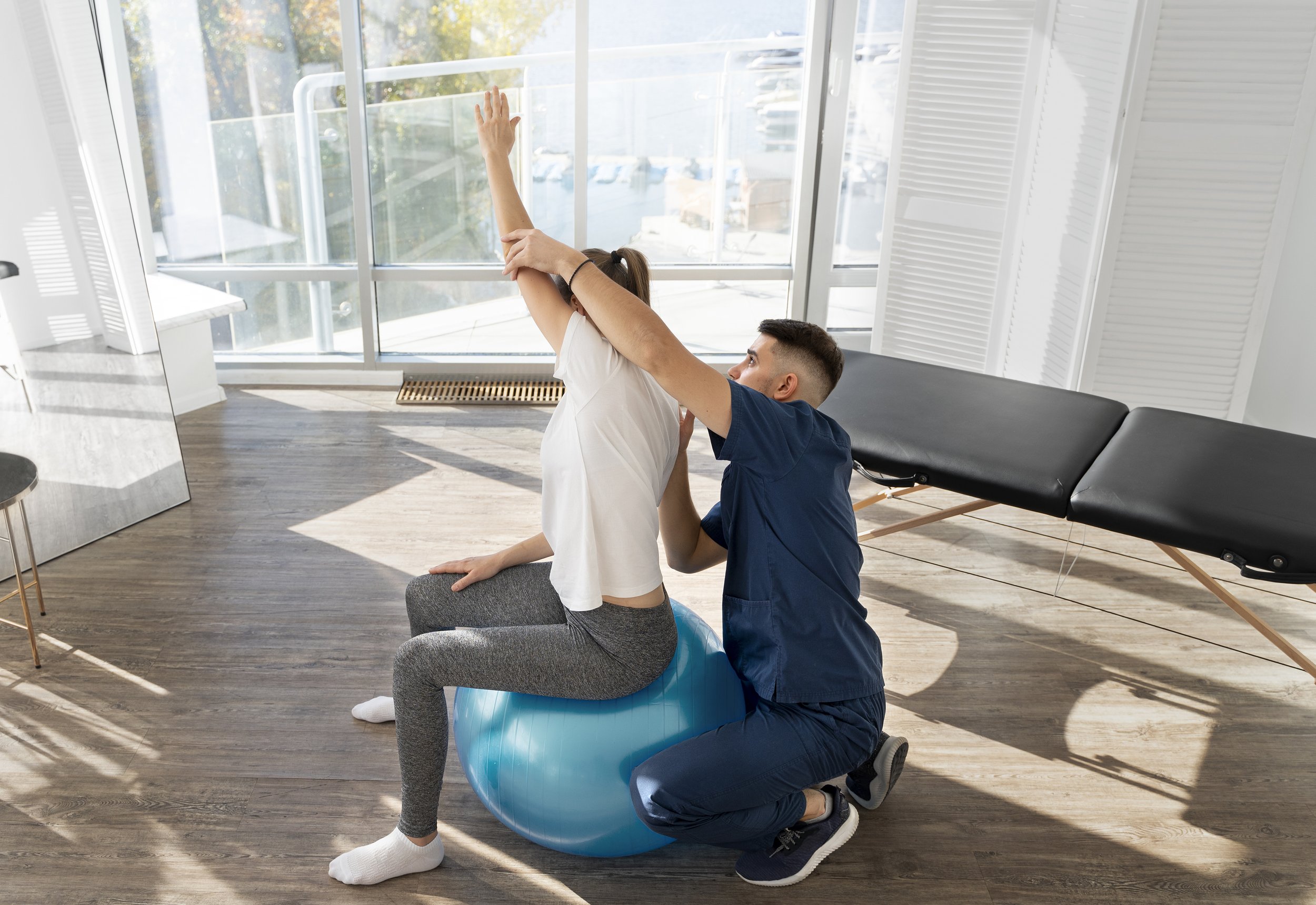 A physical therapist assists a woman performing exercises on a balance ball in a well-lit therapy room.