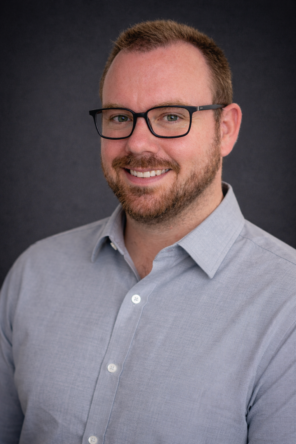 A smiling man with glasses, short light brown hair, and a beard, wearing a light gray button-up shirt against a dark gray background.