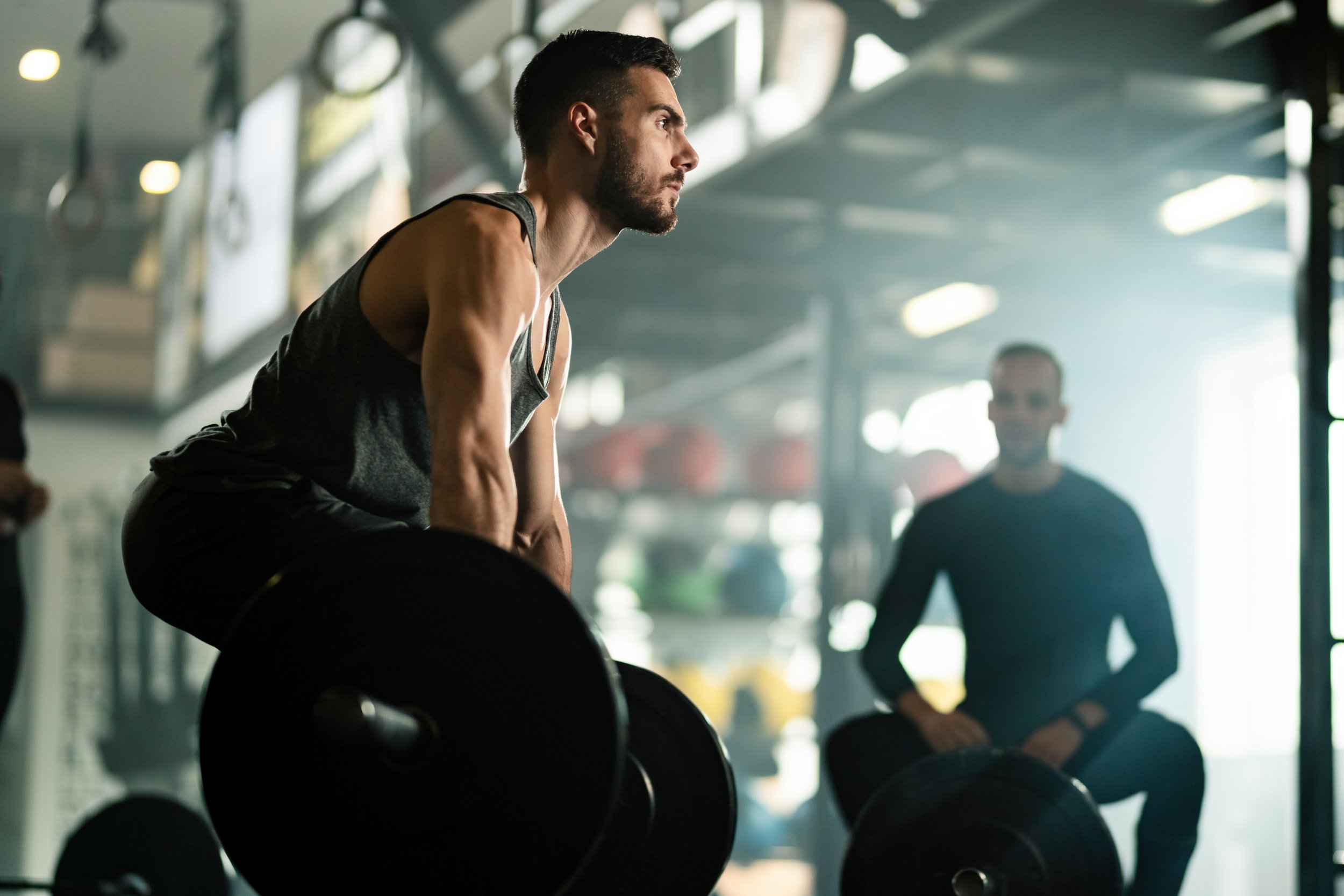A man lifting a barbell in a gym, with another person sitting nearby. The gym has a modern, industrial design with equipment and bright lighting.