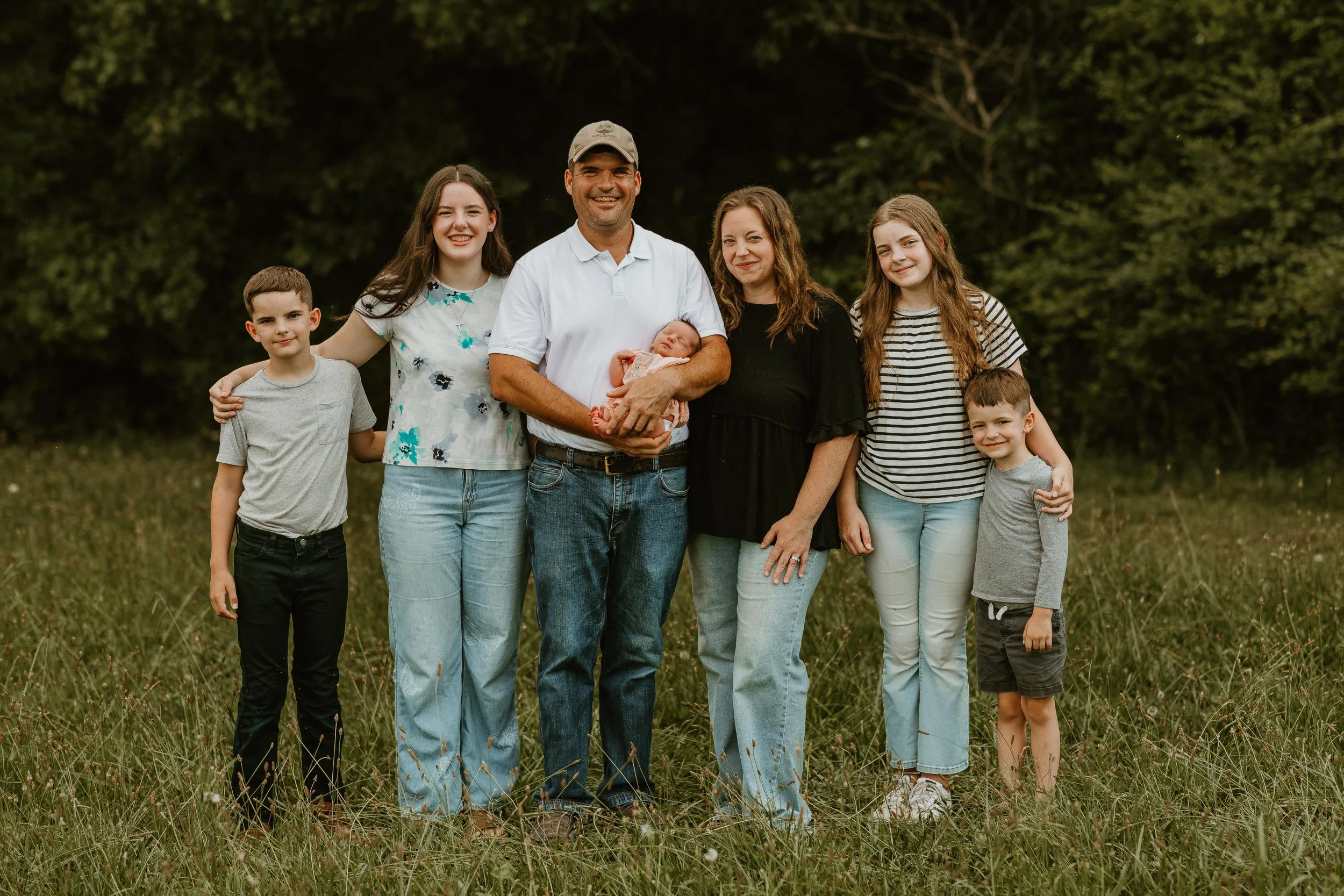 Family of seven standing outdoors on grass, smiling, with a wooded background. The group includes two adult women, one adult man holding a newborn baby, and four children of various ages.