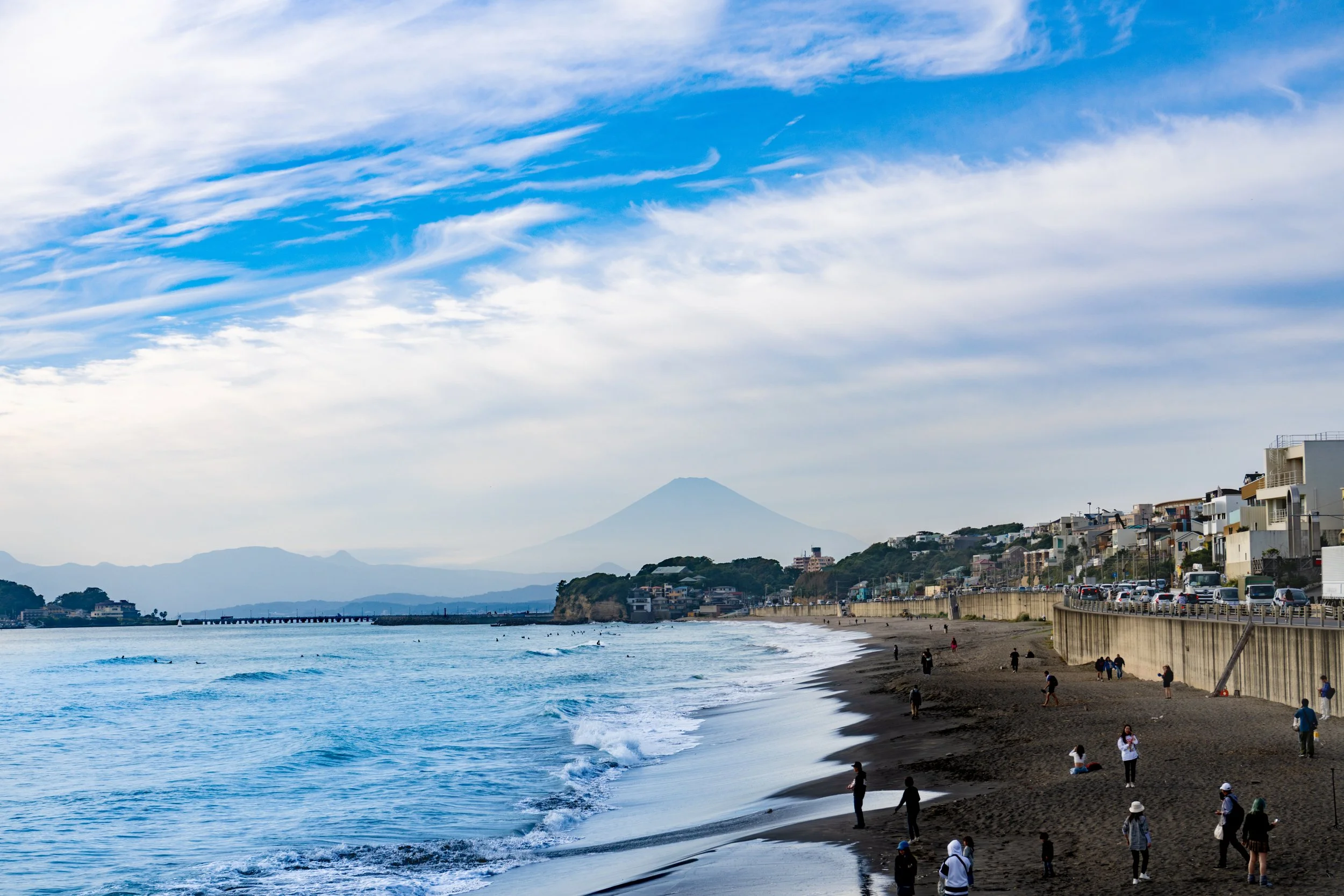 A beach scene with people walking and sitting on the sandy shore, a coastal town with houses and buildings along the hillside, and Mount Fuji in the background under a partly cloudy sky.