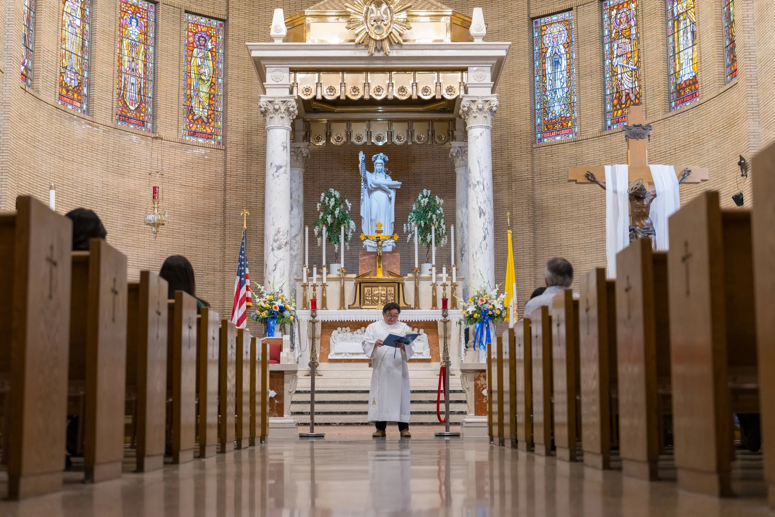 A priest reading during a religious service inside a church with stained glass windows, an altar with candles, flowers, and statues of religious figures, including Jesus on the cross.