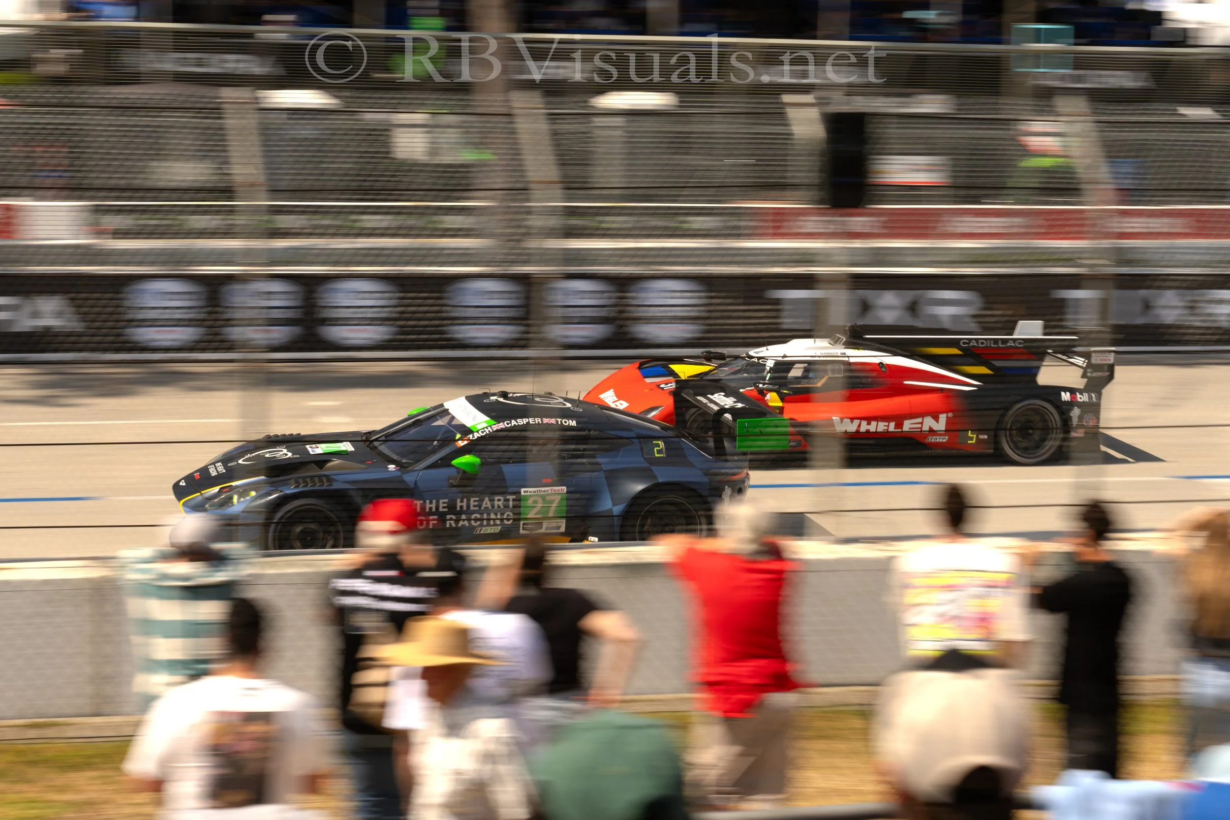 Two race cars competing on a track, with spectators watching from behind a fence. One car is black with green accents and the other is red, black, and white. Blurry motion indicates high speed.