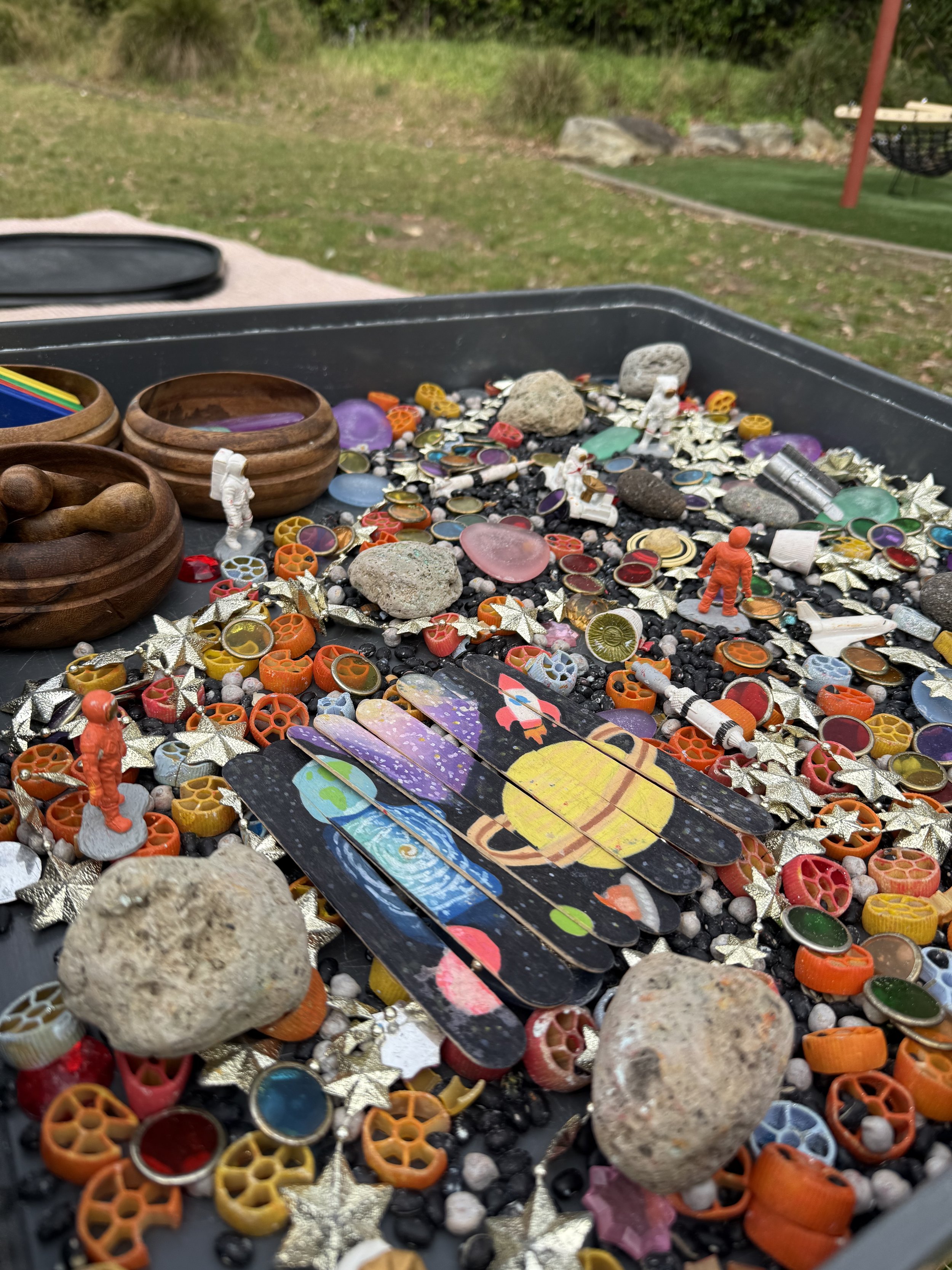 Close-up of a tray filled with small colorful objects, rocks, mini astronaut figurines, space-themed playing cards, and decorative stars, outdoors in a park.