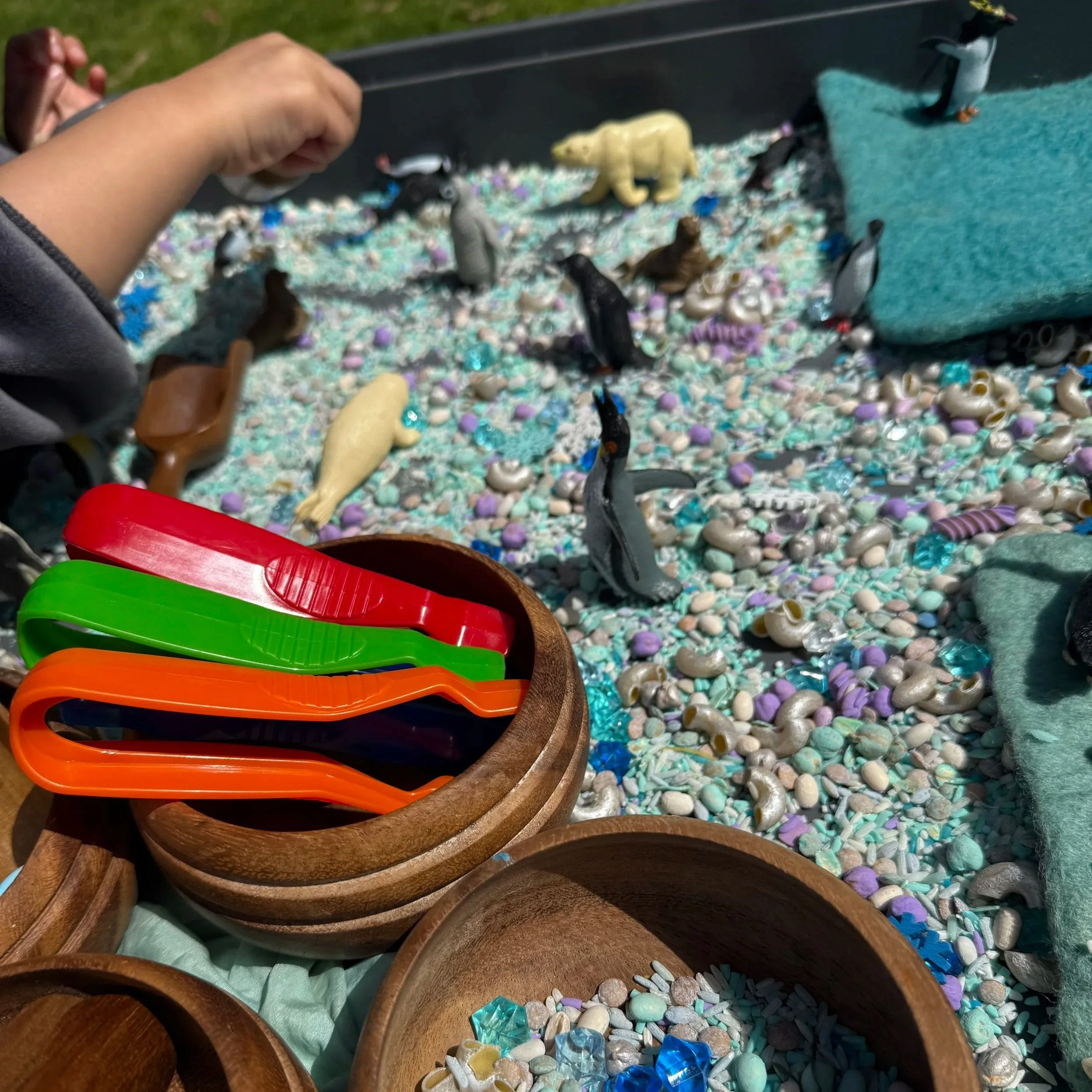 Close-up of a sensory play setup with small animal figurines on a bed of multicolored beads, shells, and glass pieces, with bowls containing colorful plastic utensils and additional small objects, and a hand reaching into the scene.