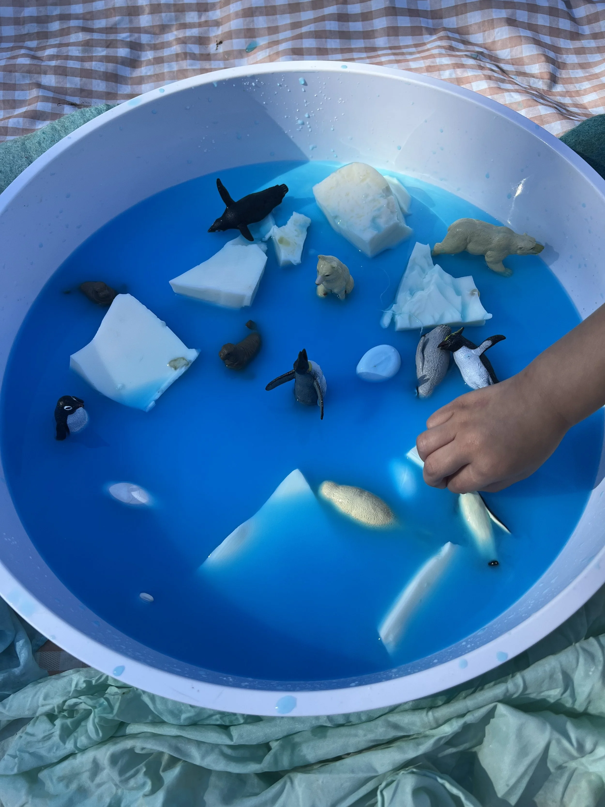 A fishing game at a fair with a round blue pool containing toy fish and blocks, with a child's hand reaching into the water to catch a toy fish.