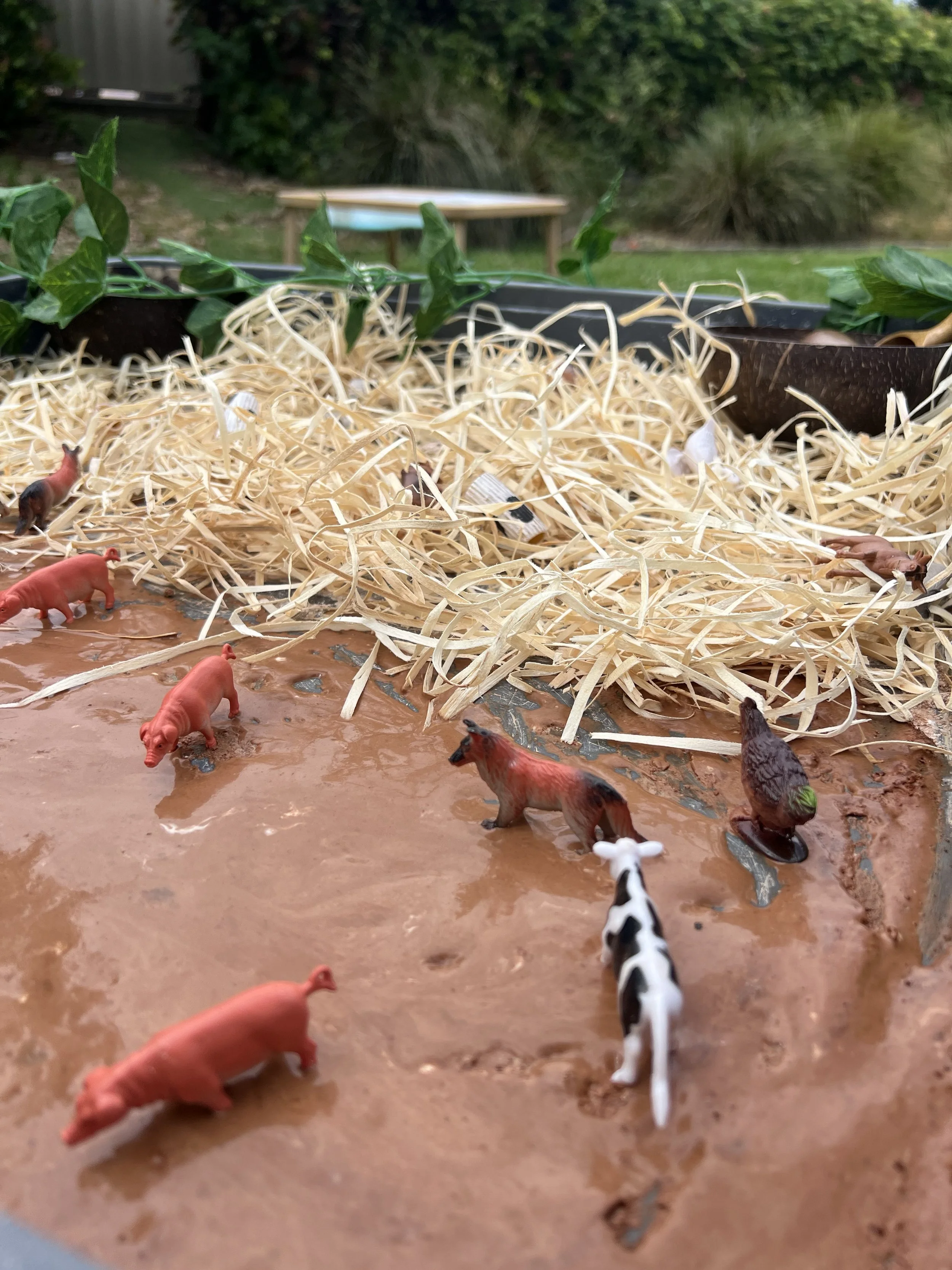 Miniature plastic animals on a muddy surface, including pigs, a horse, a cow, and an eagle, surrounded by shredded paper and green plants, with a blurred outdoor background.