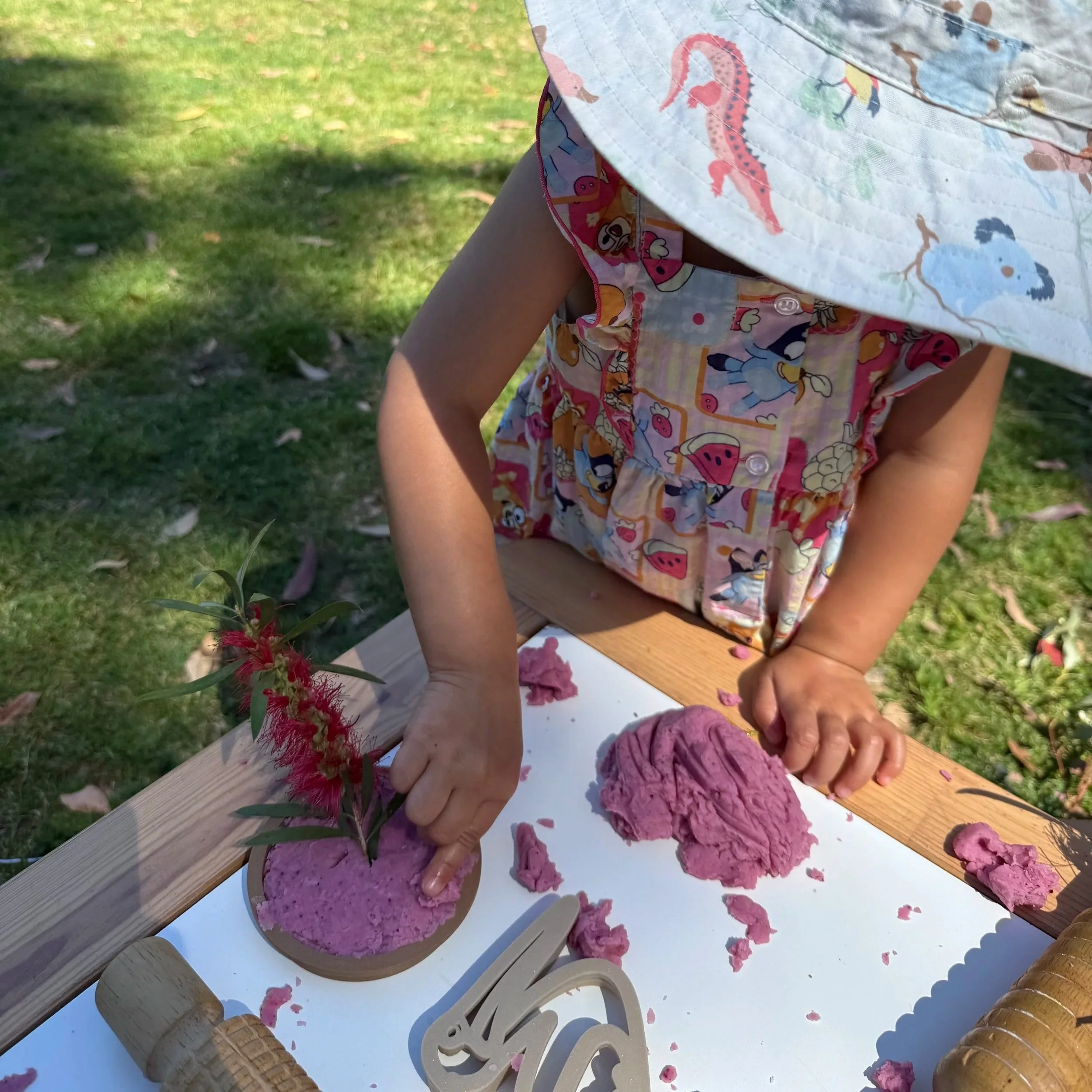 Child wearing a colorful hat and dress playing with pink playdough on a table outdoors, with a small plant nearby.