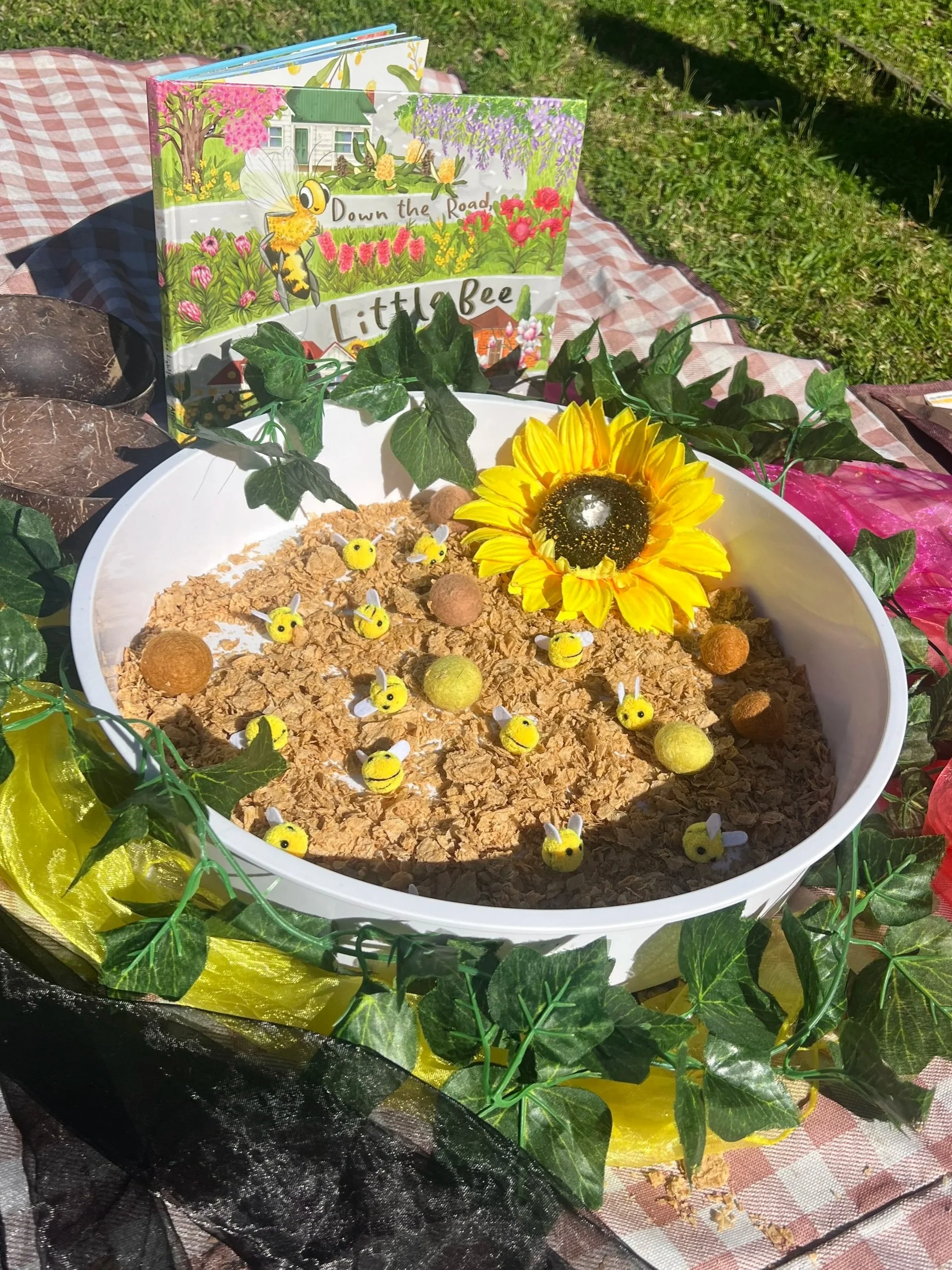 A display with small bee and round objects placed on a bed of oats inside a white dish. A sunflower decorates the side of the dish, surrounded by green artificial ivy and yellow and black fabric. In the background, there's a colorful book titled 'Down the Road, Little Bee' with illustrations of a bee, flowers, trees, and a house, set on a grassy surface.