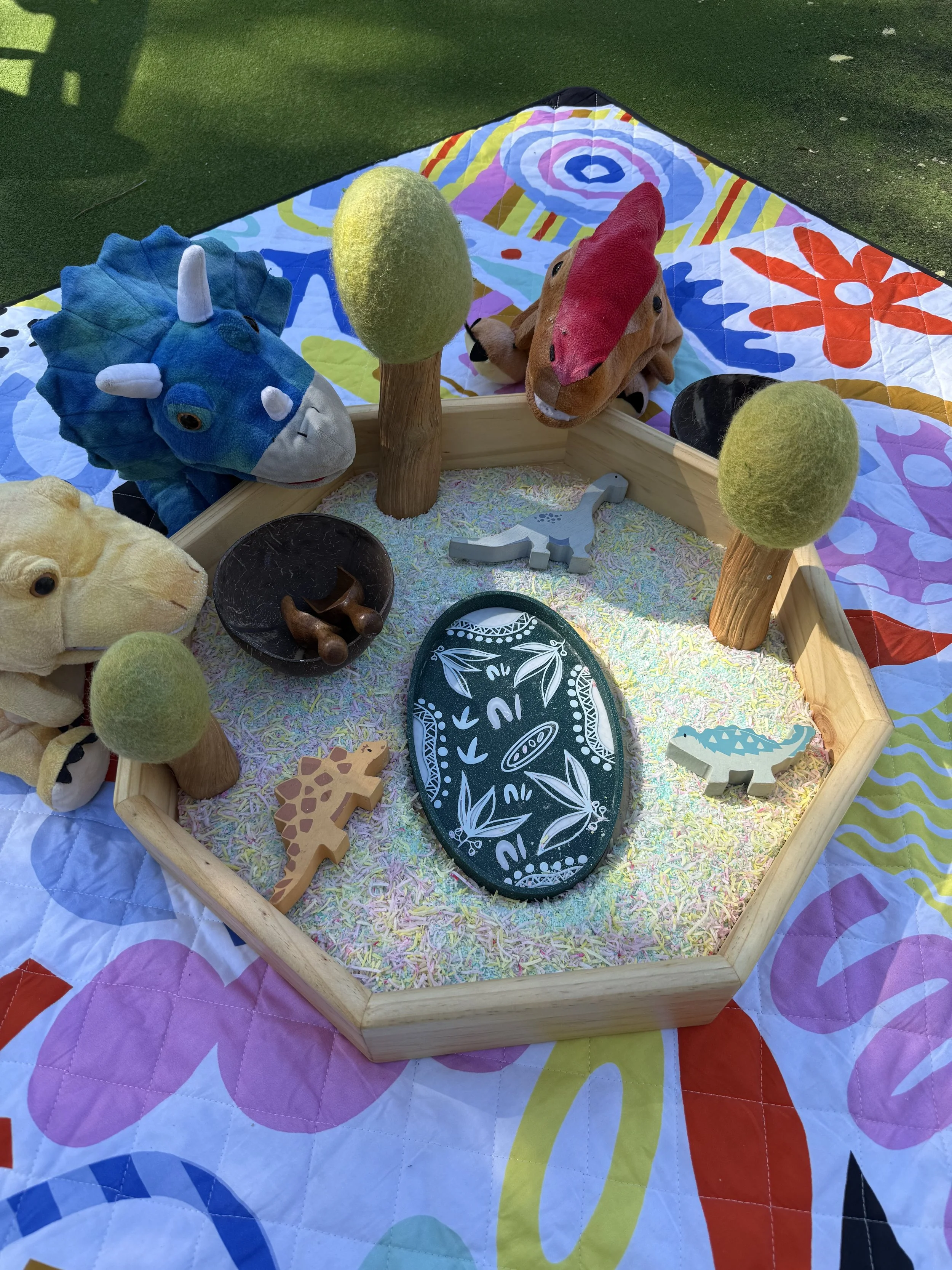 Colorful toy diorama featuring plush dinosaur and stuffed animals, wooden trees, and a painted oval centerpiece on a bed of sprinkles, all on a multicolored quilt.