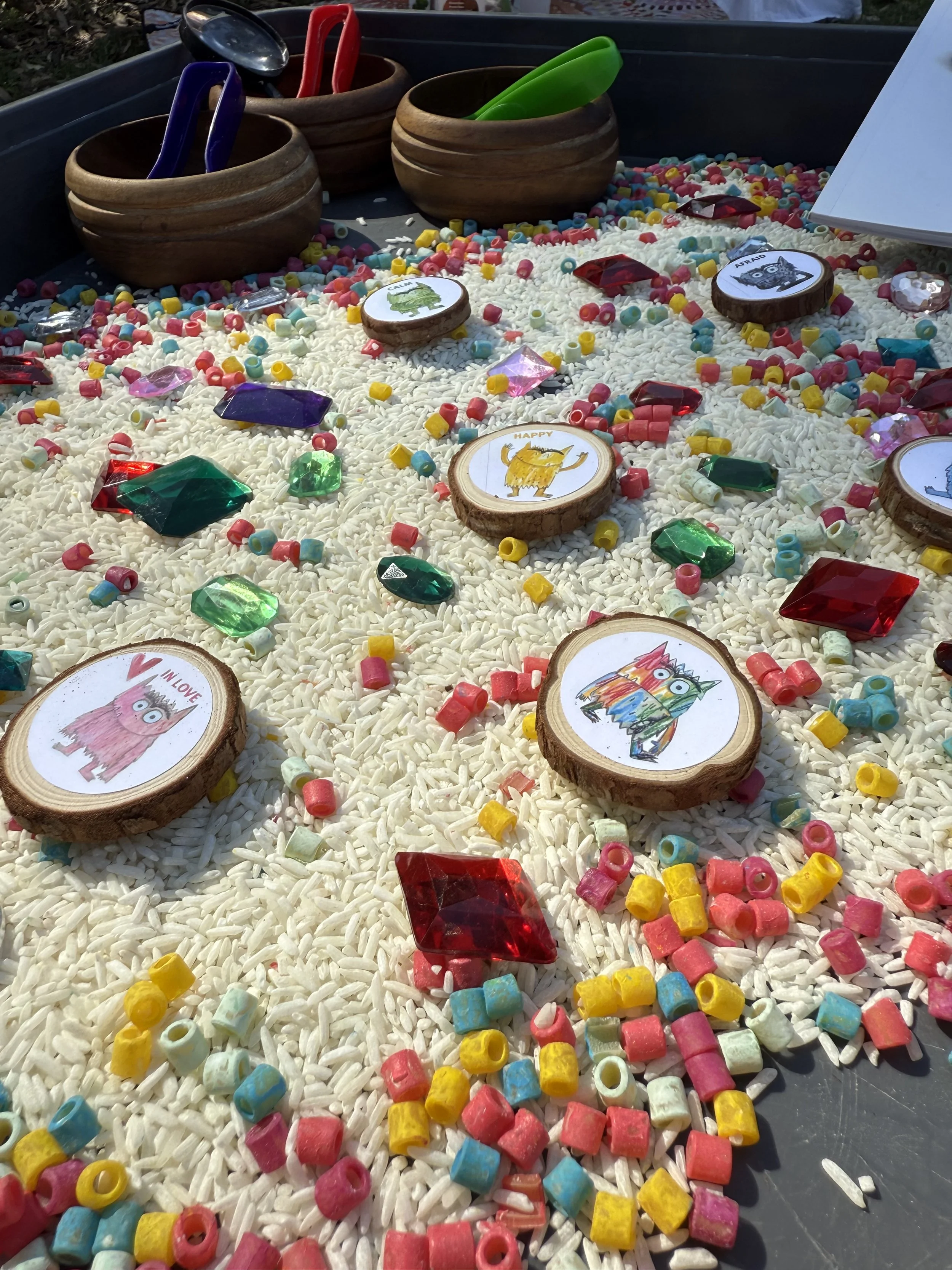 Colorful beads, wooden discs with animal illustrations, and small plastic containers on a bed of rice inside a tray, resembling a craft or game activity.