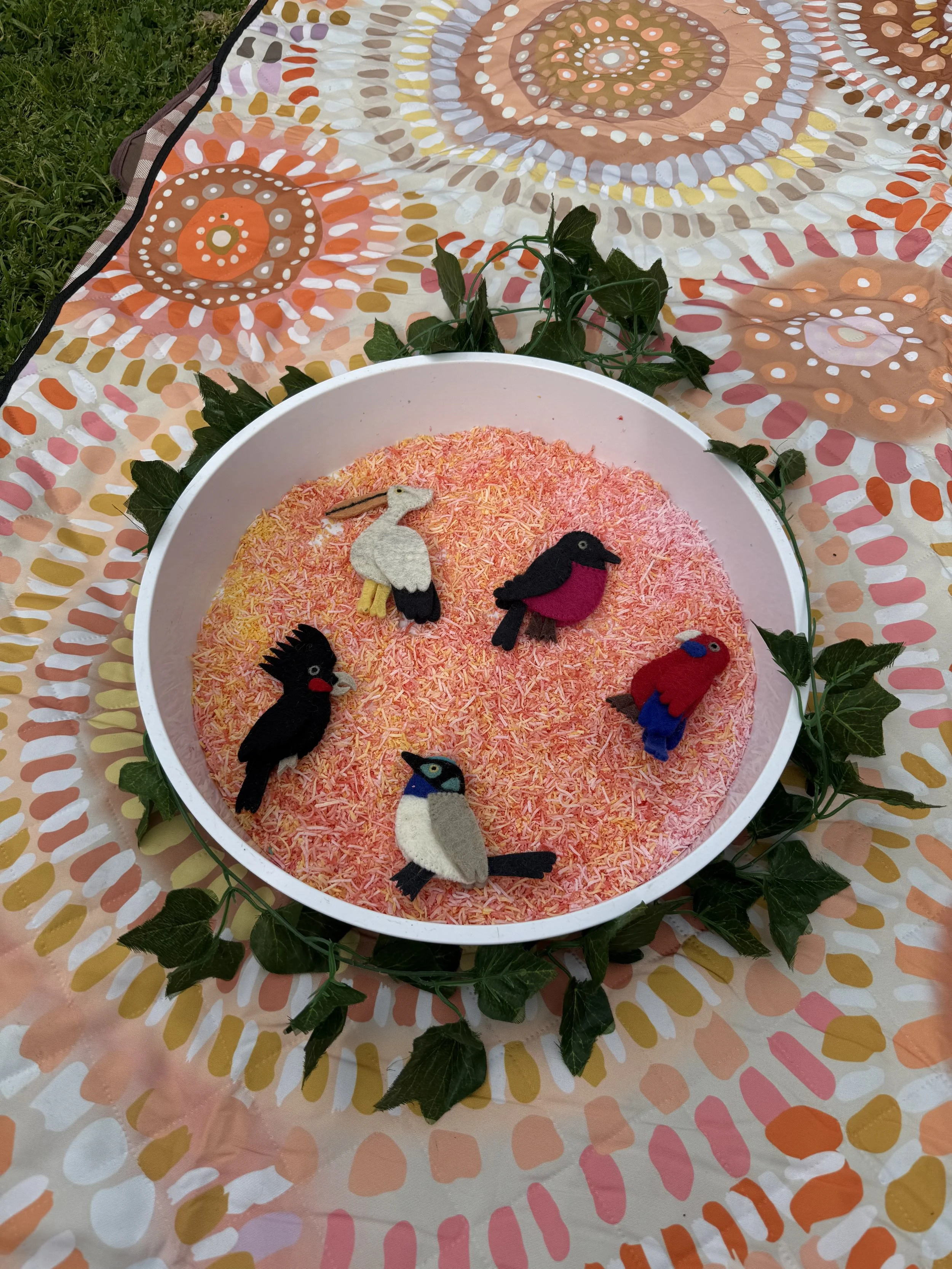 A white bowl filled with pink rice and decorative felt bird-shaped ornaments, placed on a colorful patterned cloth with a leafy green garland around it.