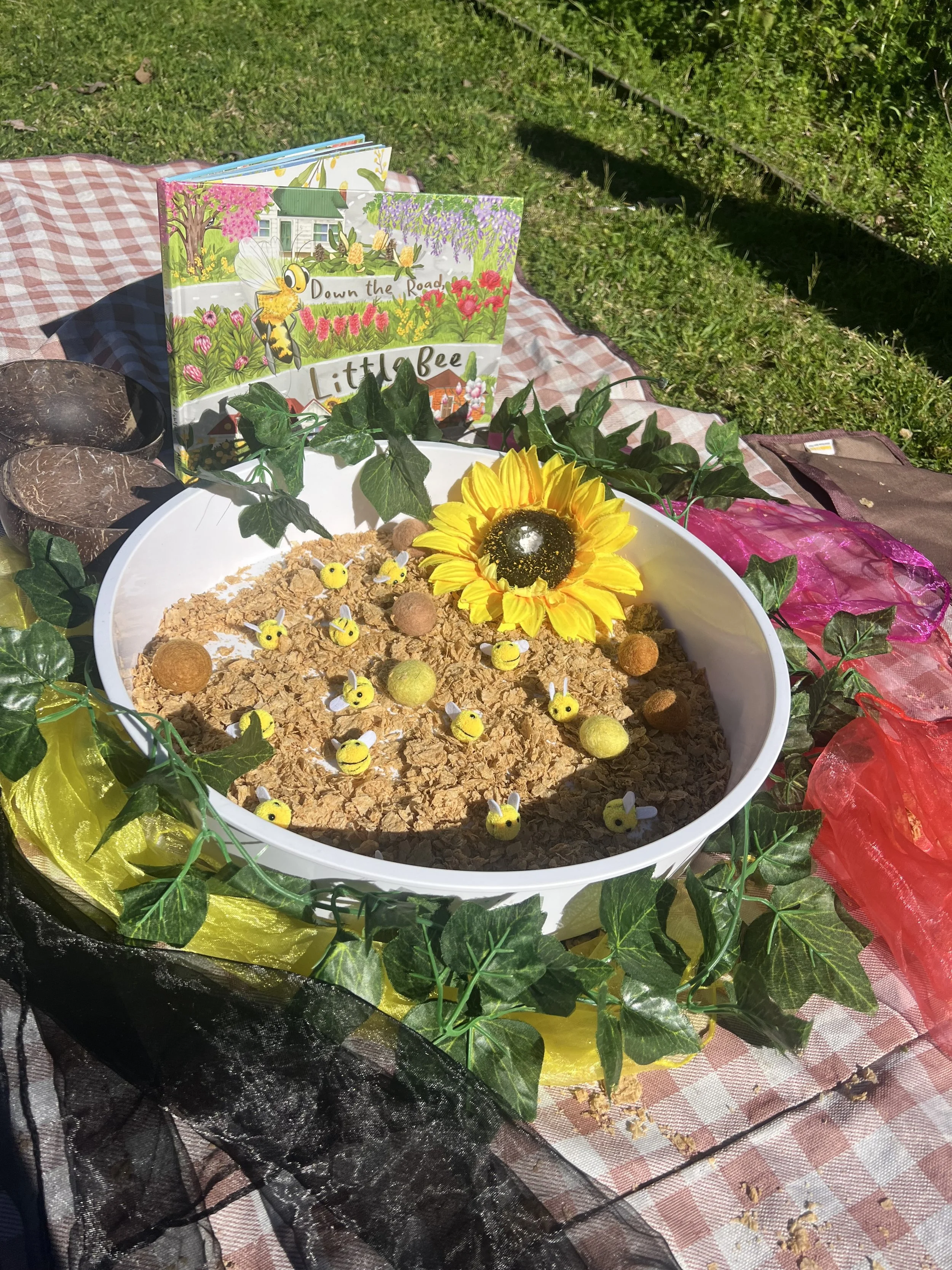 A sensory bin filled with cornflakes, small yellow bee toys, and round objects, decorated with a sunflower and green ivy, with a pink, yellow, and red fabric border, placed outdoors on a checkered picnic blanket.
