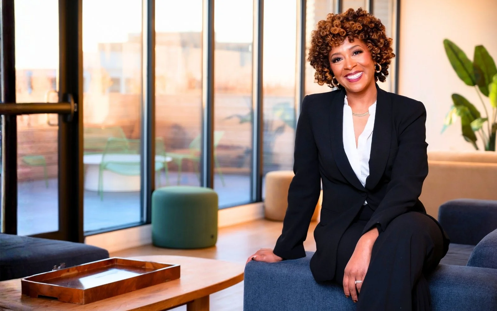 Curly haired Attorney Porcha Davis dressed in a black blazer and white shirt, smiling while sitting on a blue sofa in a modern room with large windows and a potted plant.