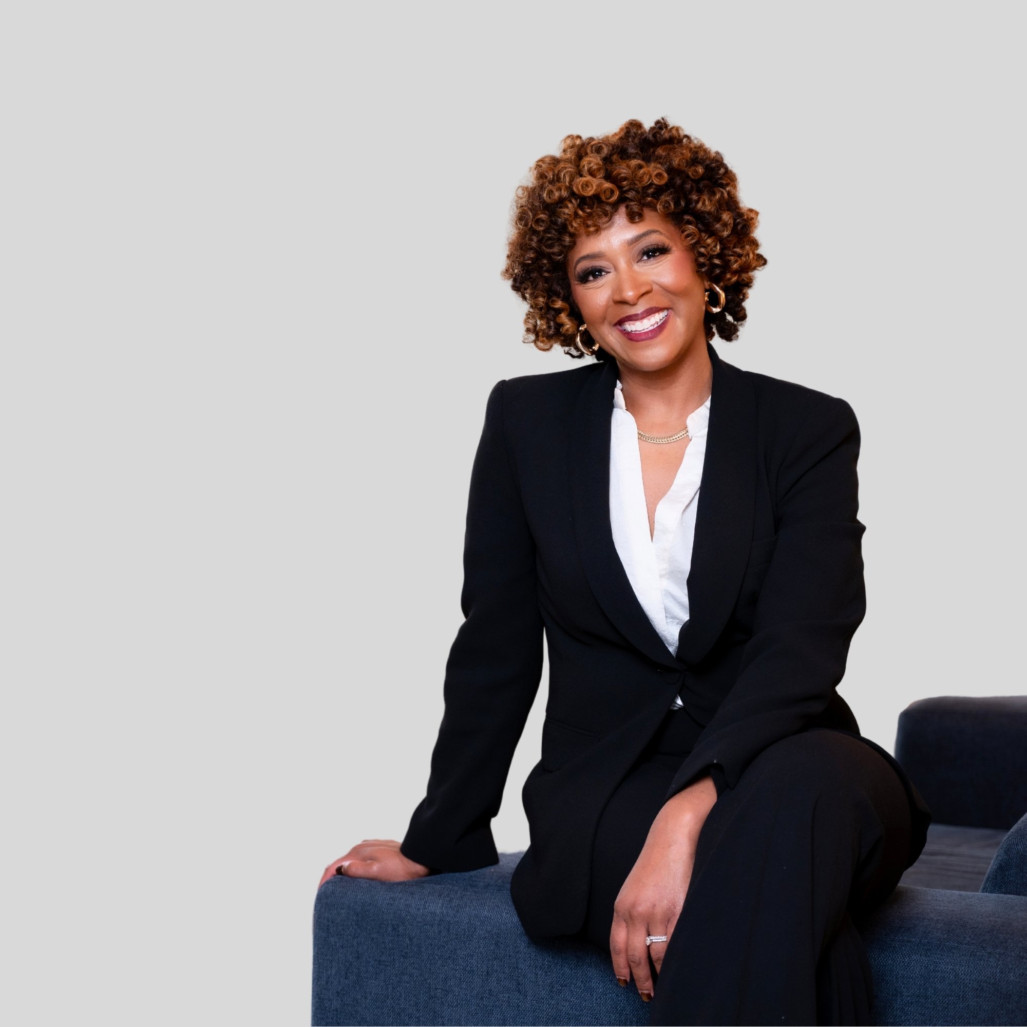 A woman with short curly brown hair wearing a black blazer, white blouse, and gold jewelry, sitting on a blue couch against a light gray background.