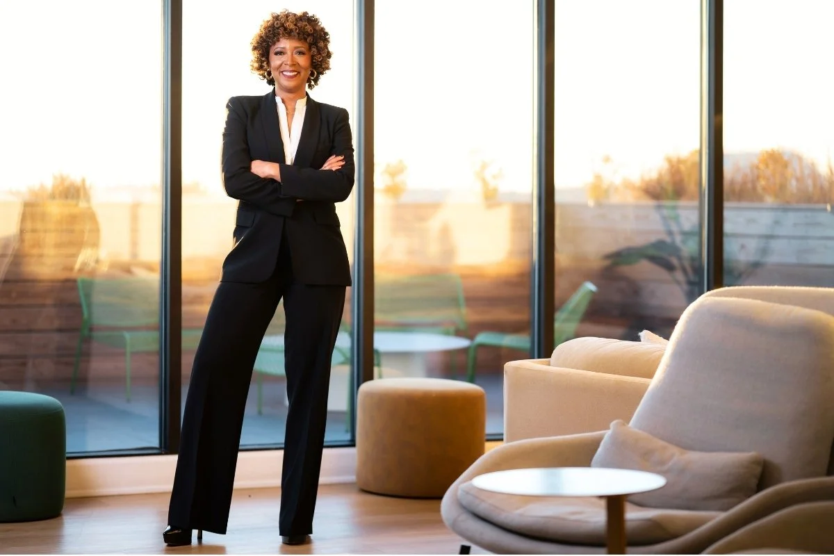 A confident woman in a black business suit standing with arms crossed inside a modern office or lounge area with large glass windows showing an outdoor patio during sunset.