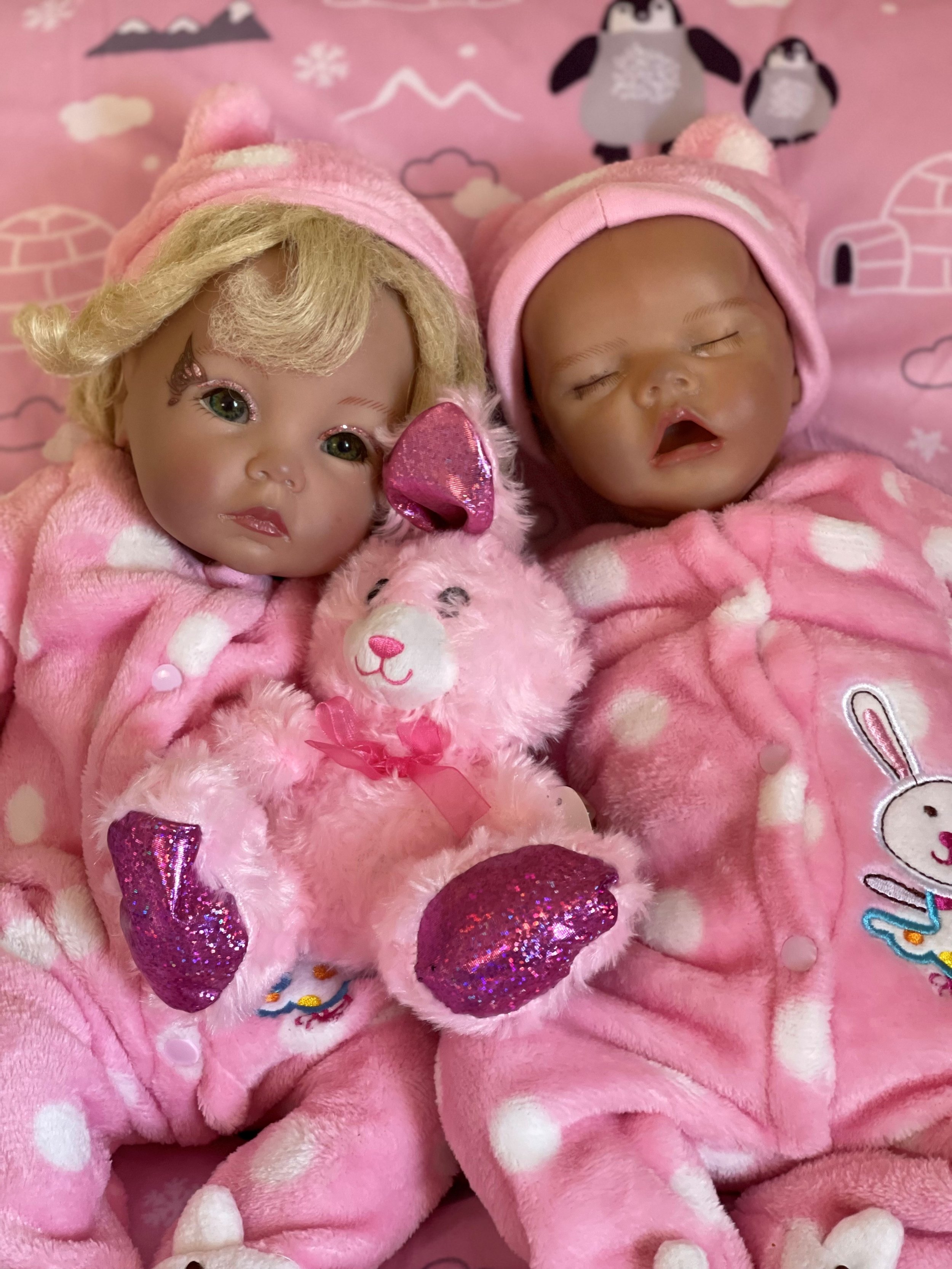 Two baby dolls dressed in pink polka dot pajamas and pink hats with pom-poms, lying side by side on a pink blanket with pandas and clouds in the background. The doll on the left has blonde hair, green eyes, and a pink sequined heart-shaped nose, hold