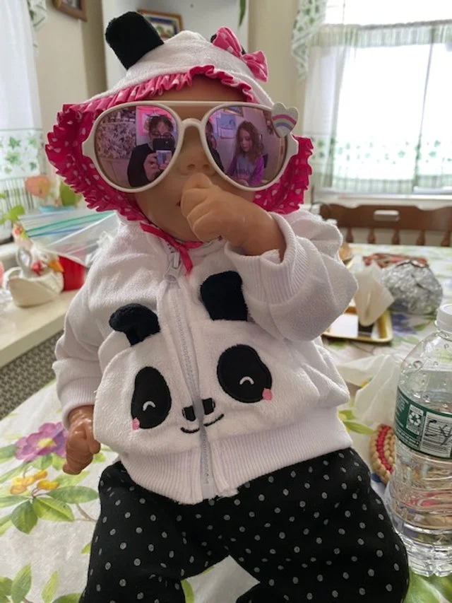 A young child wearing large pink sunglasses, a panda-themed hoodie, and a pink and white hat with a bow. The child is sitting at a table with a water bottle and other items in the background.