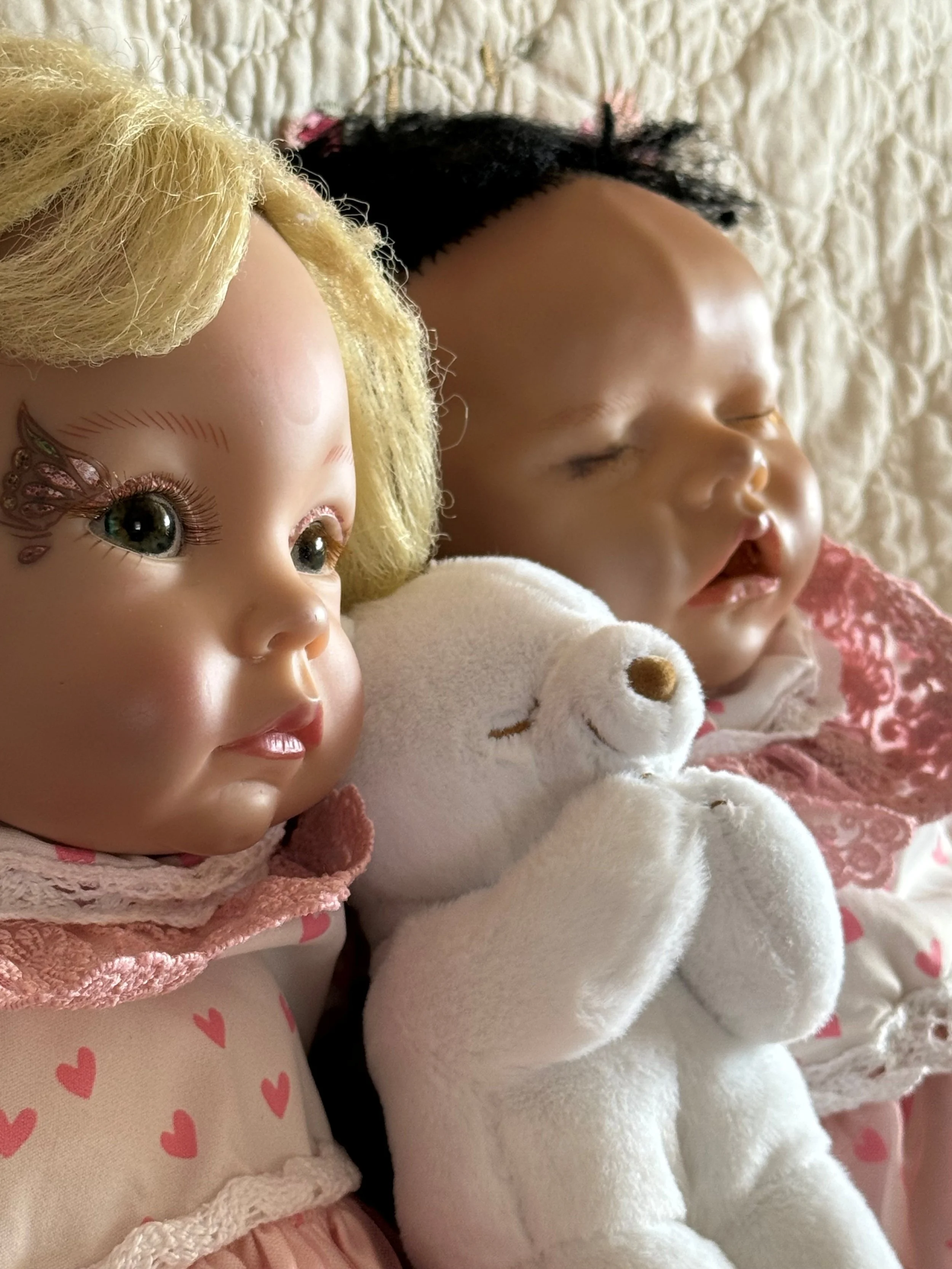 Close-up of two sleeping dolls, one with blond hair and the other with black hair, accompanied by a white teddy bear, all lying on a textured cream-colored surface.
