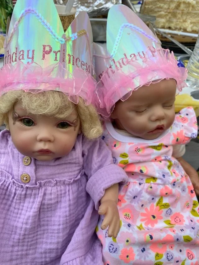 Two baby dolls wearing pink birthday princess hats with iridescent finish, decorated with pink tulle trim and 'Birthday Princess' embroidered in pink. One doll has curly blonde hair and is dressed in a purple outfit, the other has no hair and wears a