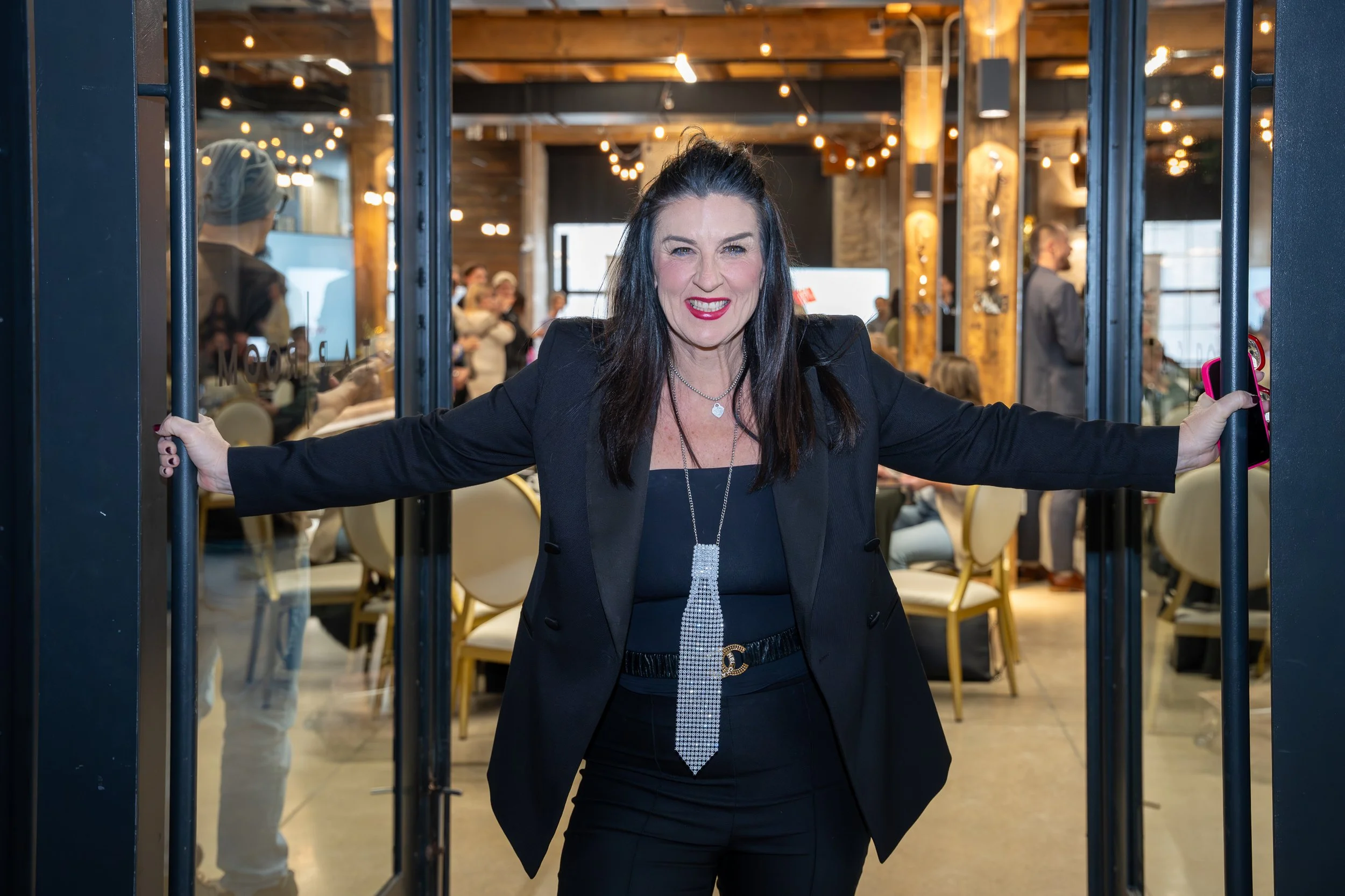 A woman with dark hair and red lipstick smiling, wearing a black blazer and pants, standing in a doorway at a social event or gathering with people and warm lighting in the background.