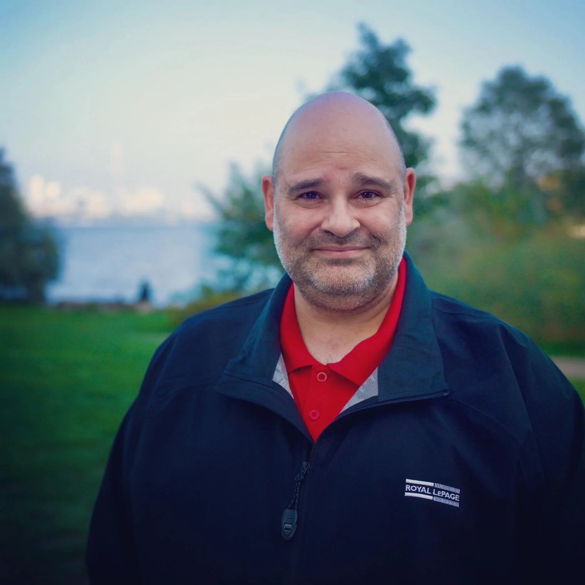 A smiling middle-aged man with a bald head and short gray beard, wearing a black Royal LePage jacket and a red polo shirt, standing outdoors in a park with trees and water in the background.
