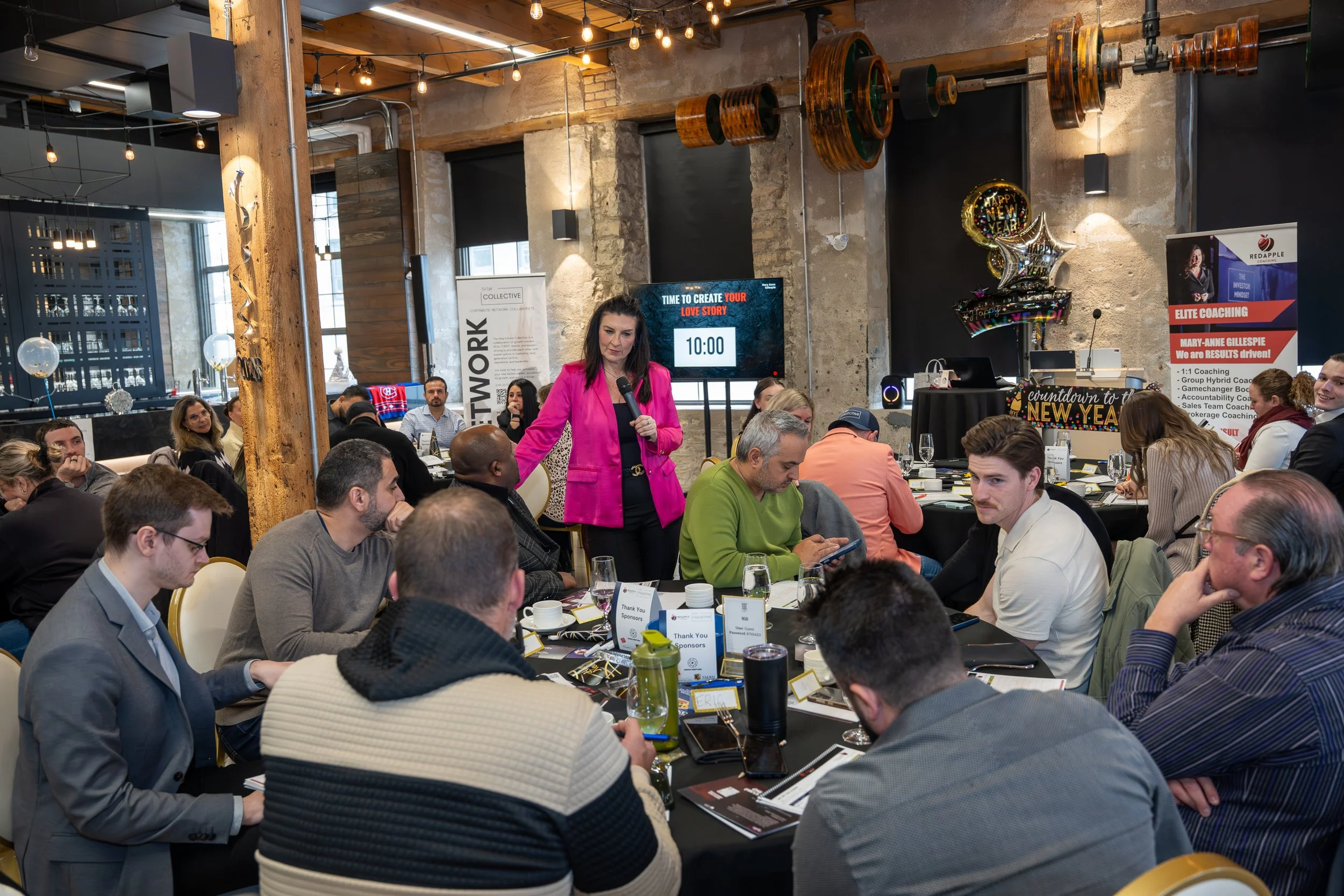 A woman in a pink blazer standing and speaking into a microphone during a business event or conference with seated attendees at tables, some using tablets or phones, in a decorated industrial-style venue with balloons, a screen displaying a timer, an