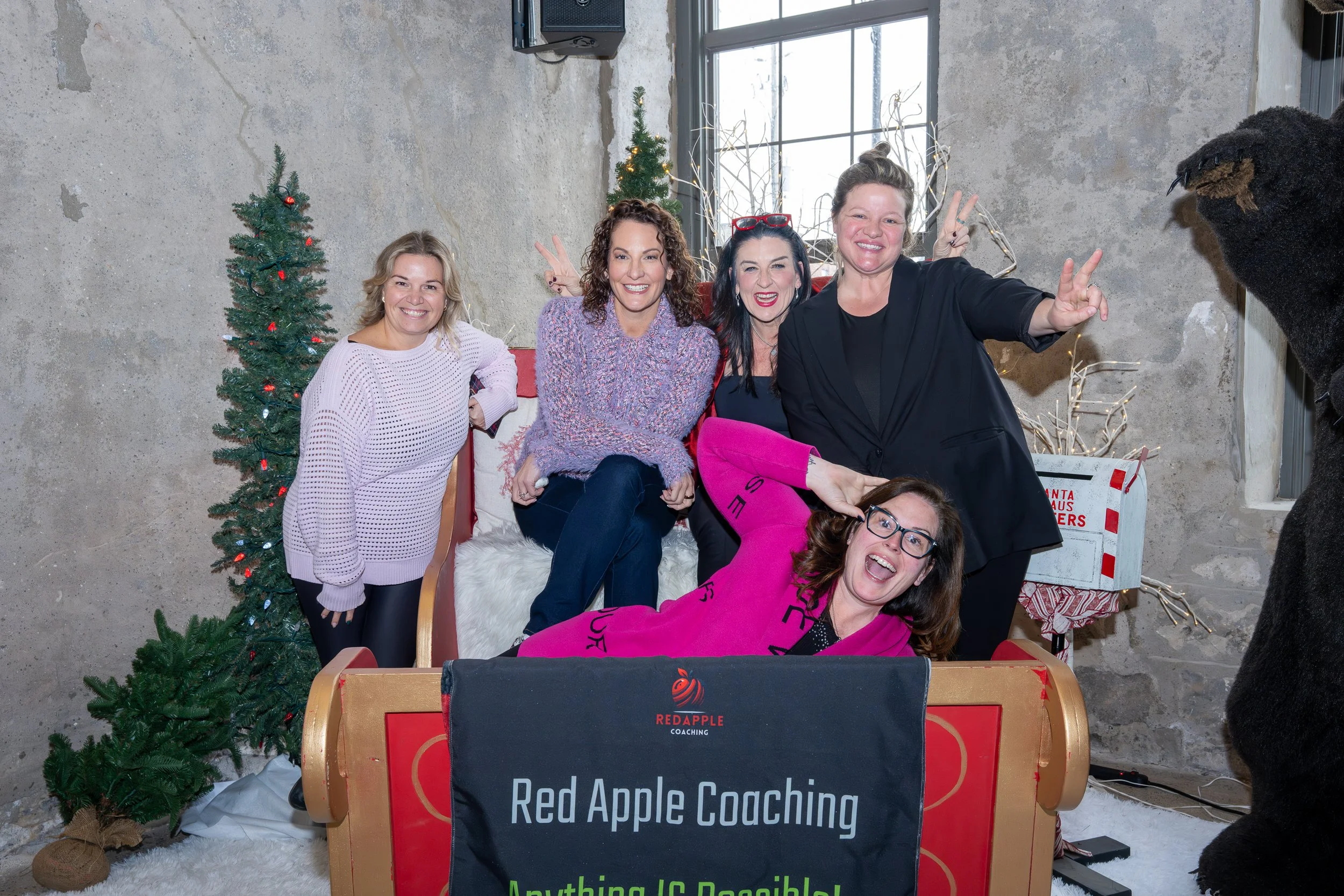 Five women sitting on a sleigh with Christmas decorations, smiling and making peace signs, with one woman lying in front wearing glasses and a pink sweatshirt, in front of a Christmas tree and holiday decorations.