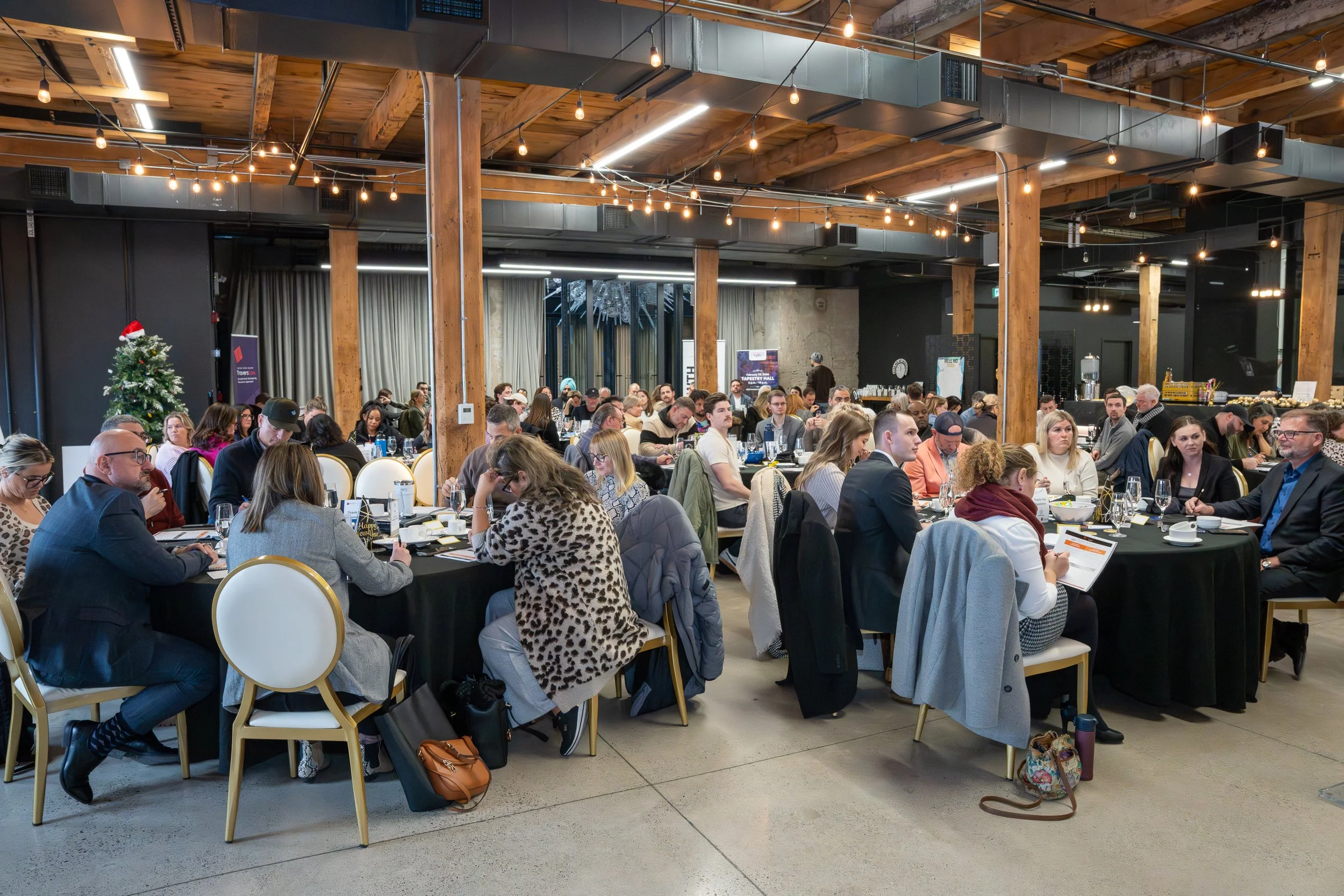 A large group of people attending a formal event or conference in a spacious venue with wooden beams and string lights, sitting at round tables covered with black tablecloths, some decorated with holiday-themed items like a Christmas tree with a Santa hat.